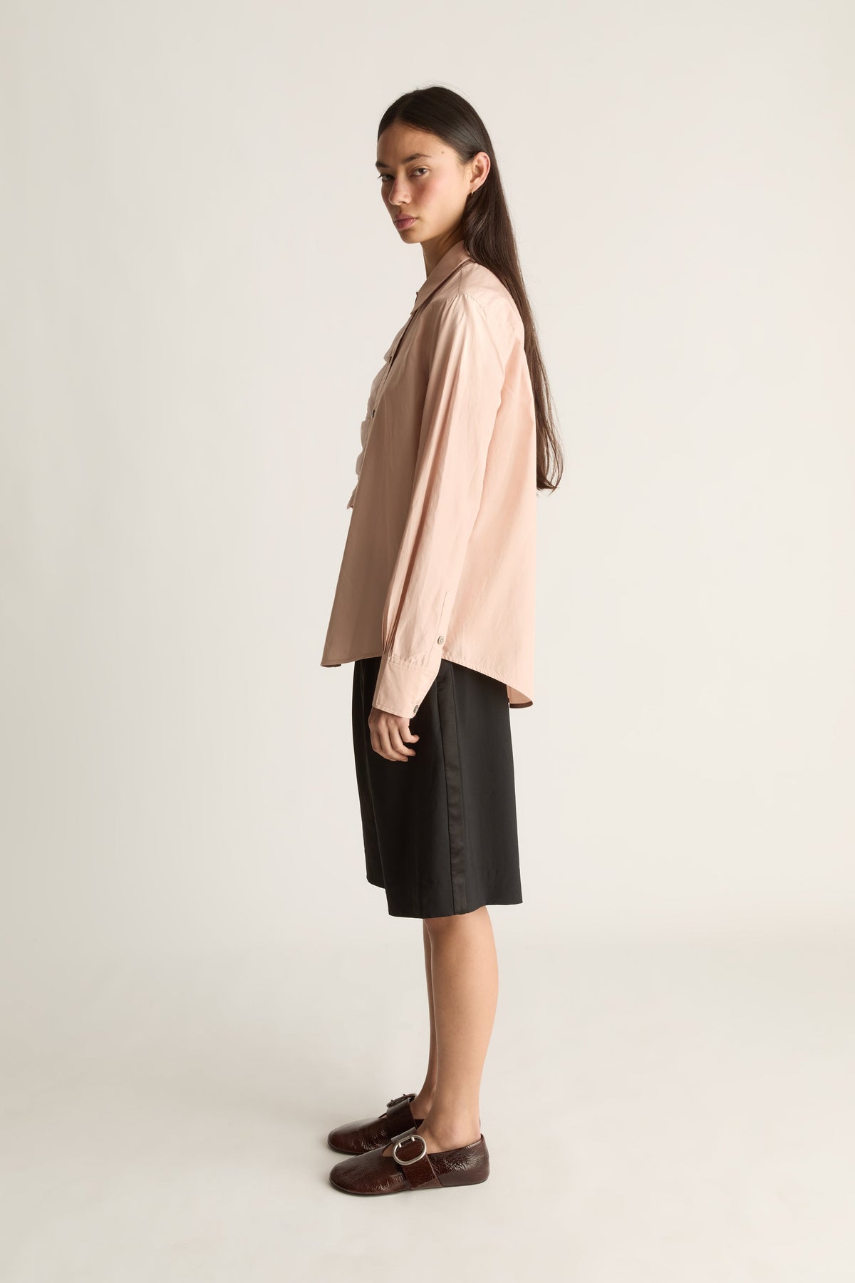A woman stands in profile wearing a light blush pink long-sleeve button-up shirt, black shorts, and dark brown loafers with a buckle strap, against a plain light beige studio background.