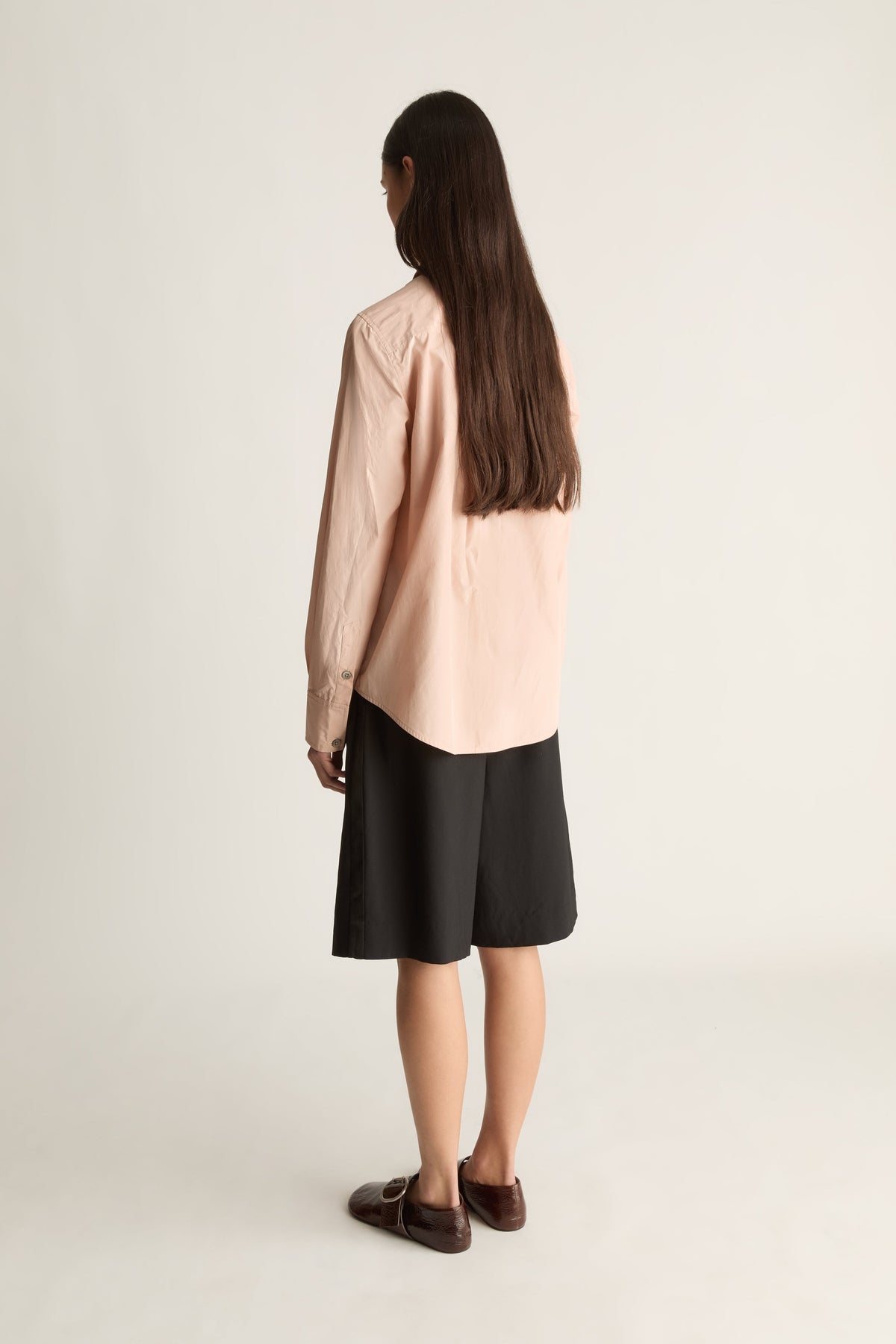 A woman is shown from behind wearing a light peach-pink long-sleeve shirt and a black midi skirt, paired with dark brown loafers, against a plain light gray studio background.