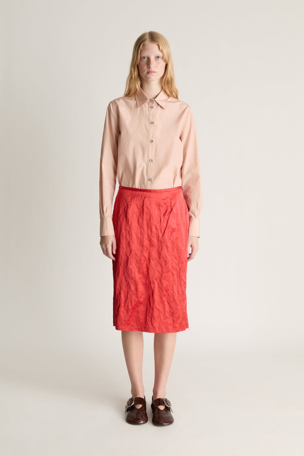 Front view of a woman wearing a pale pink button up shirt and a red silk midi skirt against a white studio background