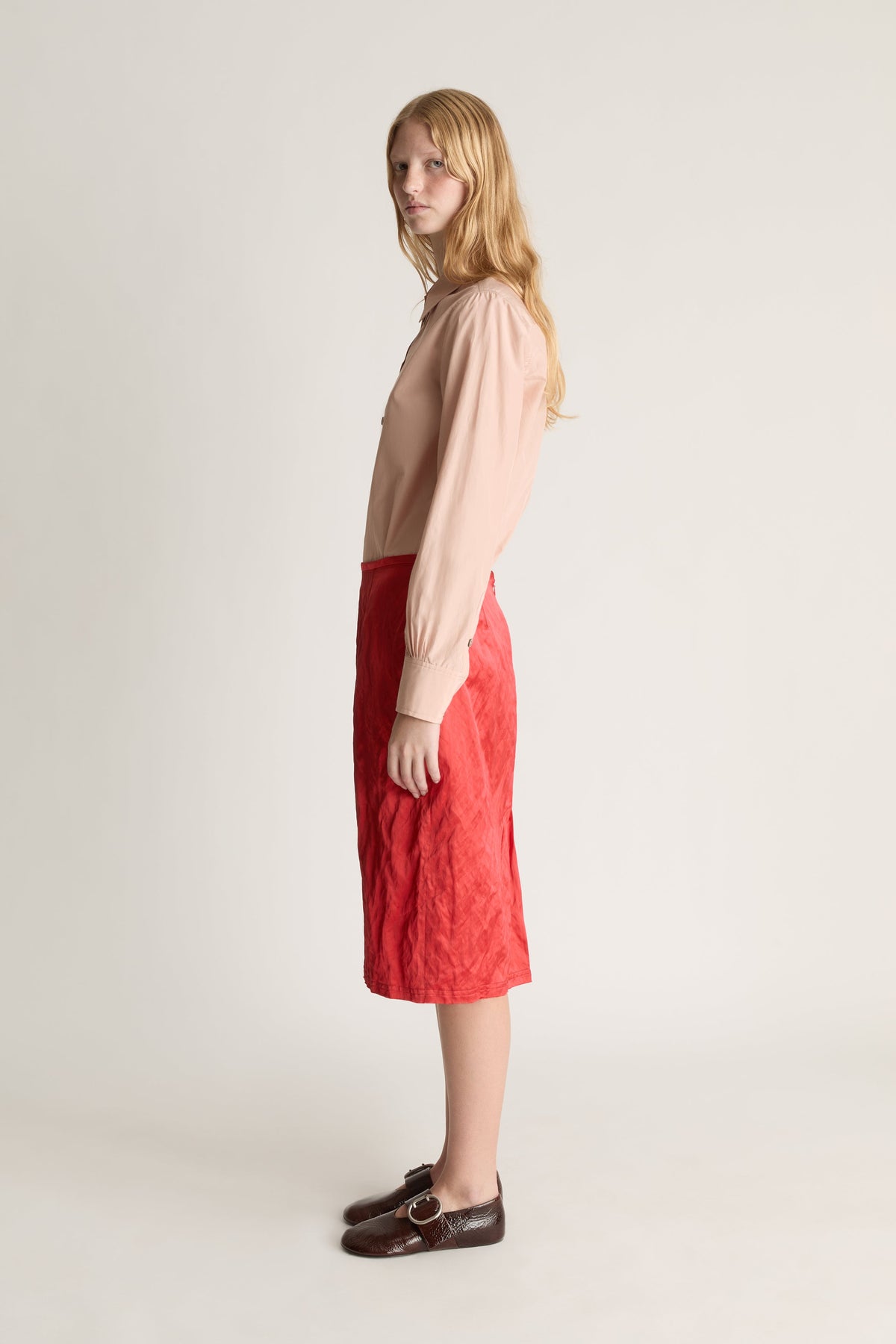 Profile view of a woman wearing a pale pink long sleeve button up shirt and a red midi skirt against a white studio background. 