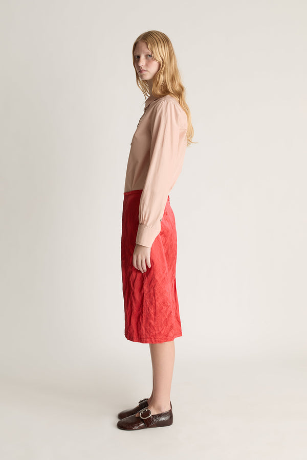 Profile view of a woman wearing a pale pink long sleeve button up shirt and a red midi skirt against a white studio background. 