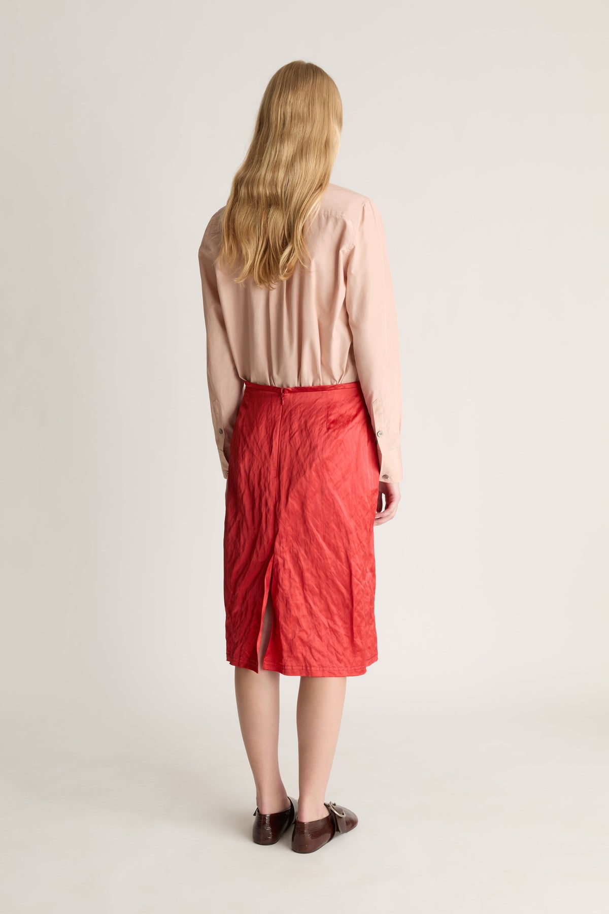 Back view of a woman wearing a pale pink button up shirt and red midi skirt against a white studio background. 