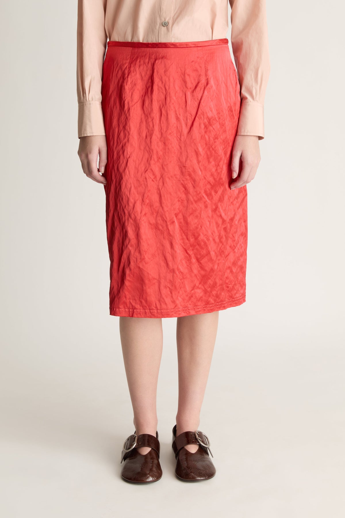 Close up of a woman wearing a pale pink shirt and red midi skit with brown shoes against a white studio background. 