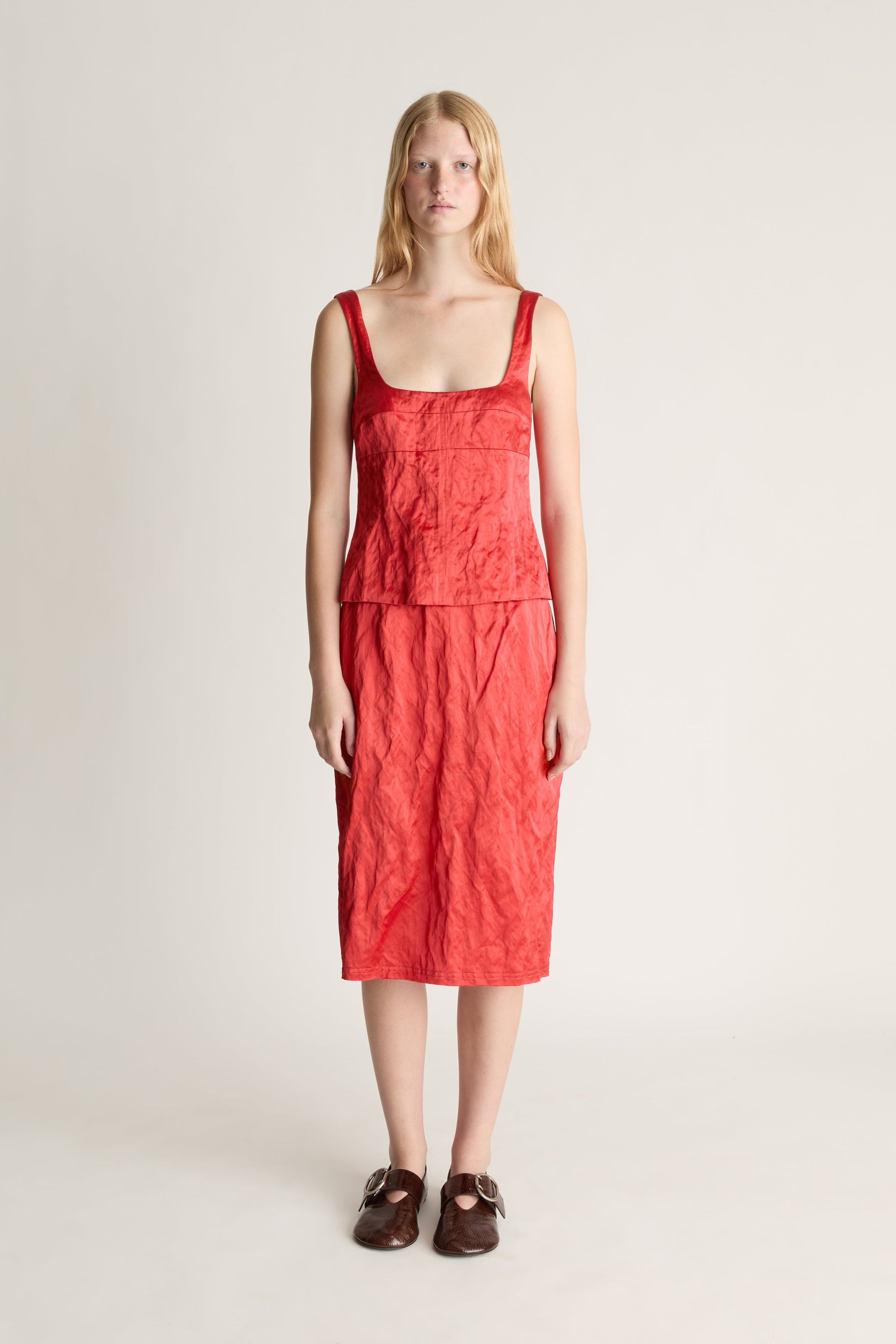 Front view of a woman wearing a red silk cami and midi skirt with brown shoes against a white studio background. 
