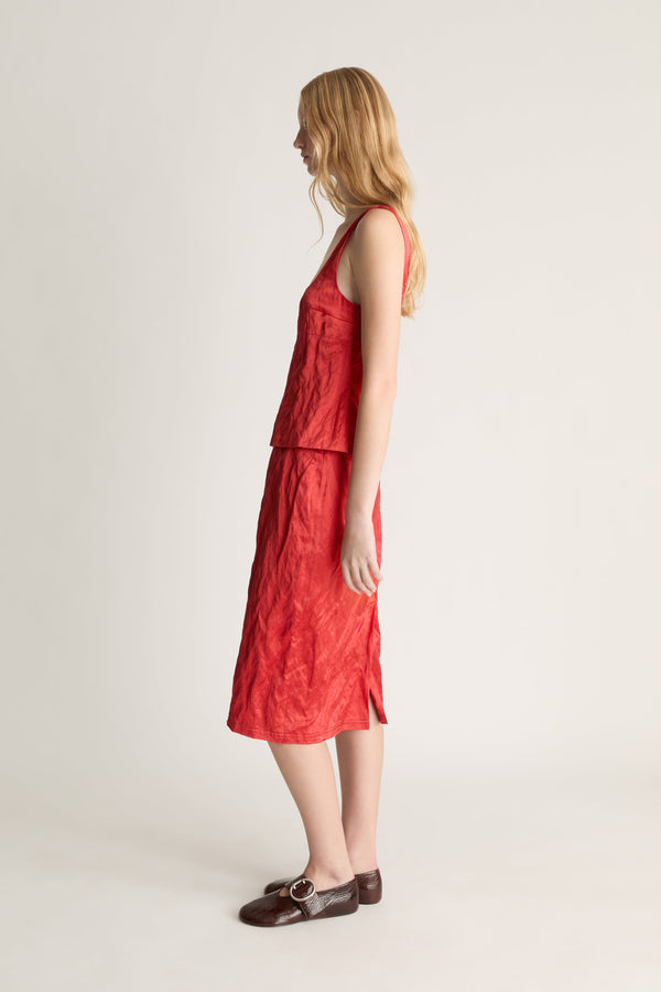 Woman standing in side profile wearing a red silk tank top and midi skirt with brown shoes against a white studio background. 