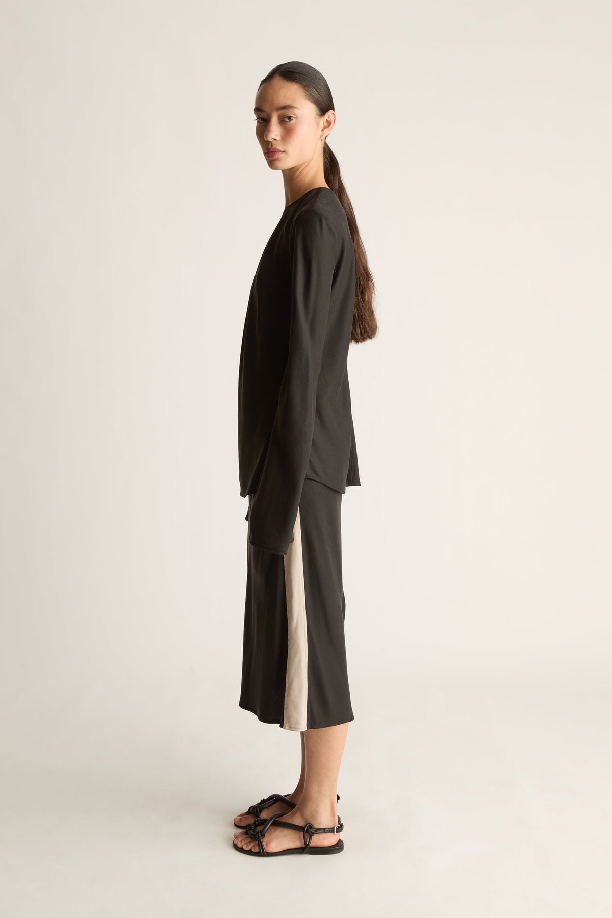A model stands in profile wearing a loose black long-sleeve top and matching black midi skirt beige panel up the sides, paired with black strappy sandals, against a plain light gray studio background.