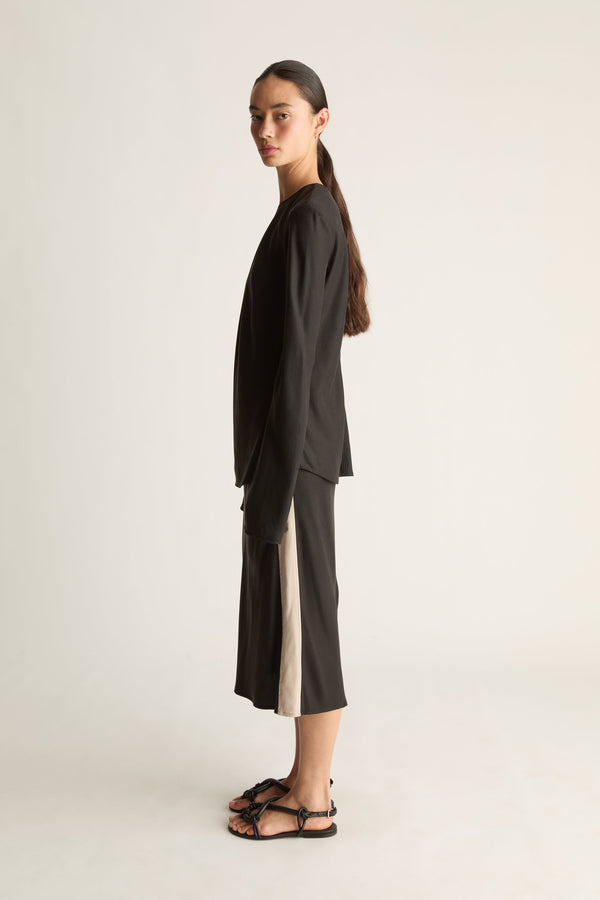A model stands in profile wearing a loose black long-sleeve top and matching black midi skirt beige panel up the sides, paired with black strappy sandals, against a plain light gray studio background.
