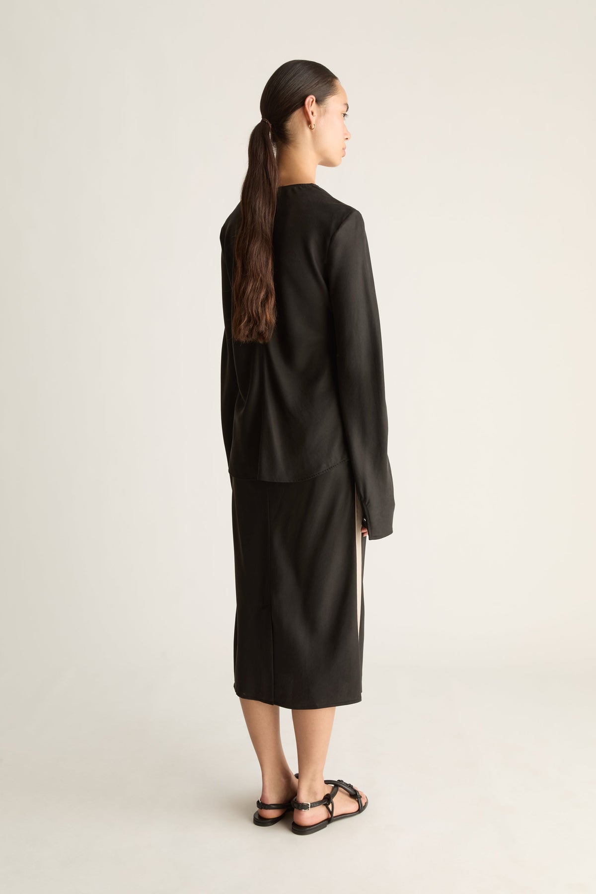 A woman stands in profile wearing a black long-sleeve top and matching midi skirt set against a plain light gray studio background.