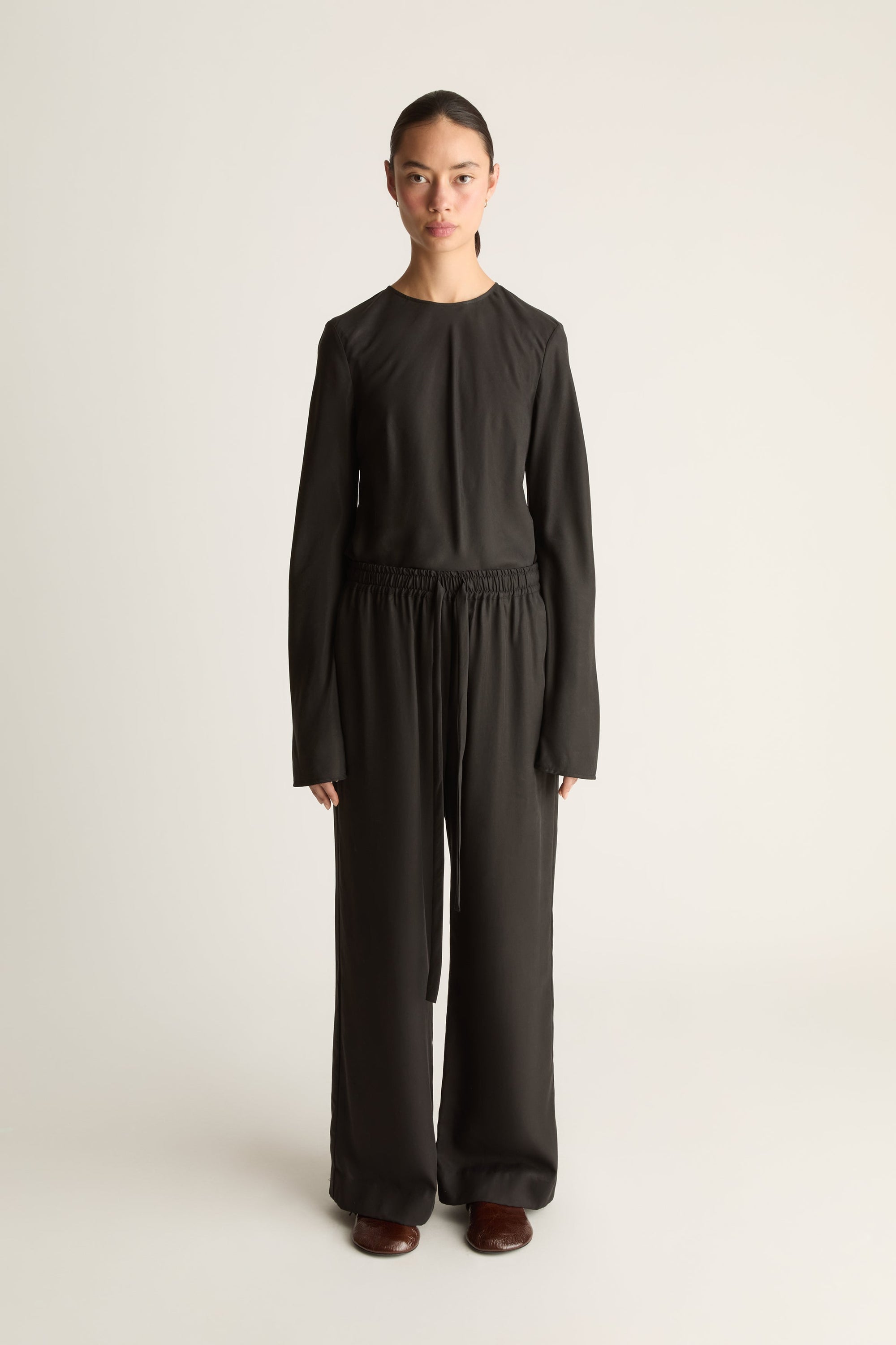 Front view of a woman wearing a black long sleeve shirt with a pair of black drawstring elasticated trousers standing against a white studio background.
