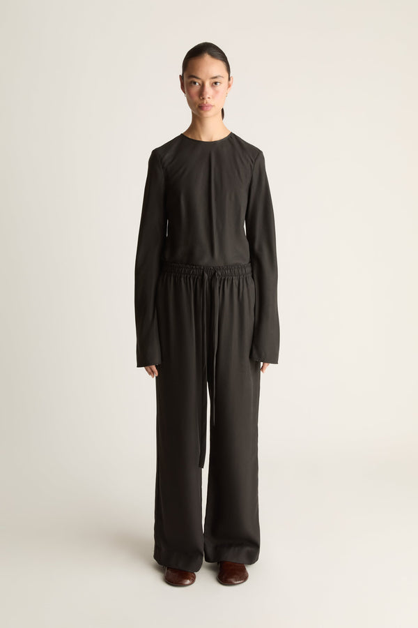 Front view of a woman wearing a black long sleeve shirt with a pair of black drawstring elasticated trousers standing against a white studio background.