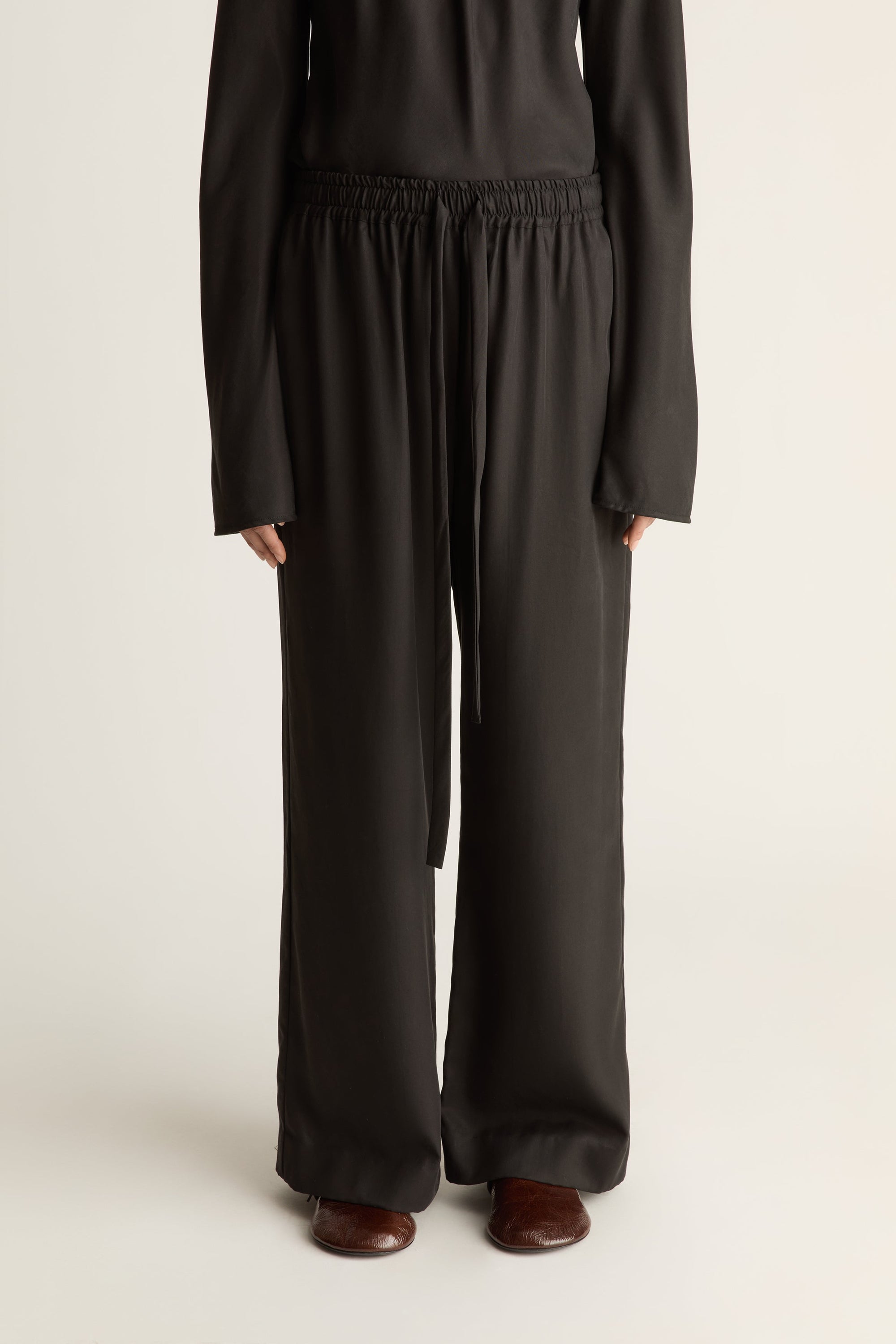A person wearing loose black wide-leg pants with an elastic waistband and drawstring, paired with a black long-sleeve top and brown shoes, photographed against a plain light background.