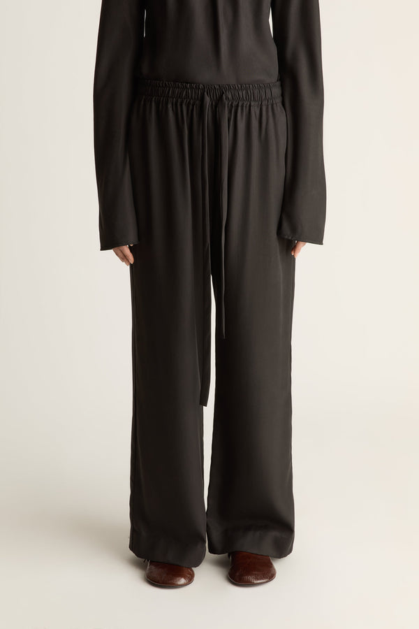A person wearing loose black wide-leg pants with an elastic waistband and drawstring, paired with a black long-sleeve top and brown shoes, photographed against a plain light background.