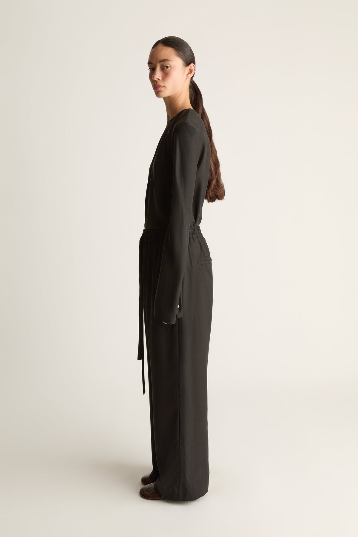 A woman stands in profile wearing a long black wrap-style jumpsuit or romper with a waist tie, photographed against a plain light studio background.