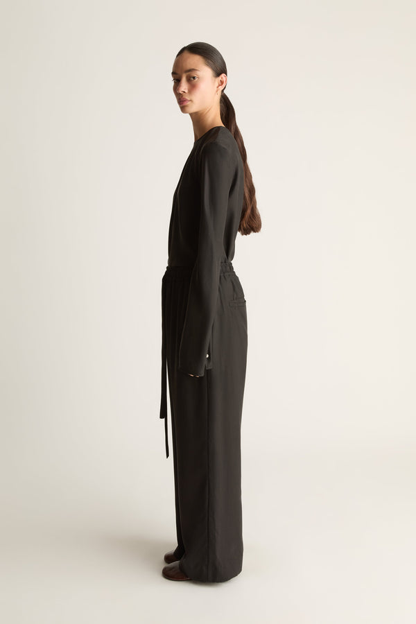 A woman stands in profile wearing a long black wrap-style jumpsuit or romper with a waist tie, photographed against a plain light studio background.