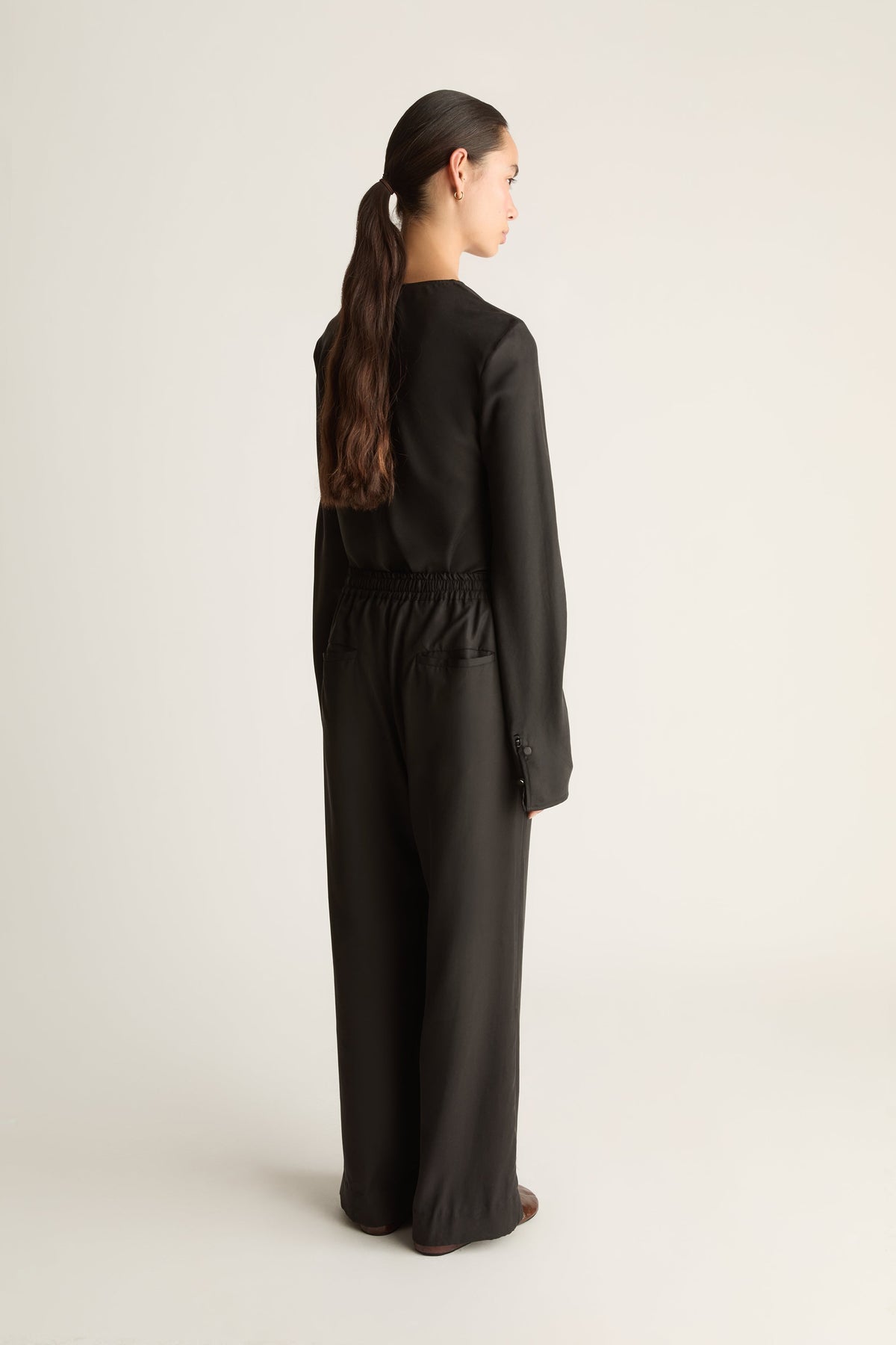 A woman with her hair in a ponytail is shown from the back wearing a black long-sleeve jumpsuit with a gathered elastic waistband, photographed against a plain light gray studio background.
