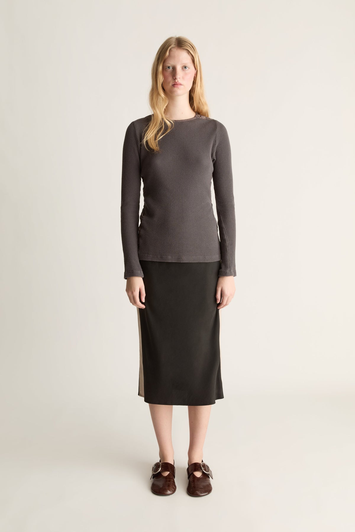 A woman stands against a plain light beige studio background wearing a dark gray long-sleeve knit top, a black midi skirt, and dark brown loafers.