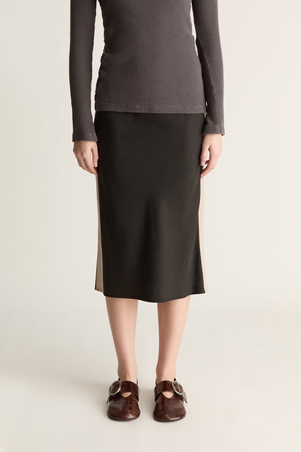 A model wearing a dark charcoal gray ribbed long-sleeve top and a black midi skirt with beige side panels, paired with brown leather Mary Jane shoes, against a light beige studio background.