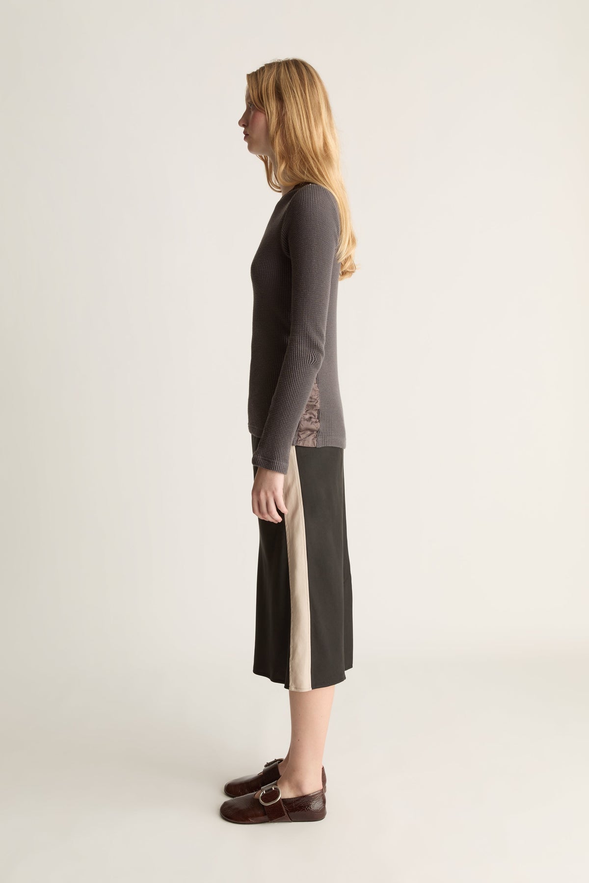 A woman stands in profile wearing a dark charcoal long-sleeve knit top and a dark midi skirt with a beige side stripe, and dark brown loafers, against a plain light background.