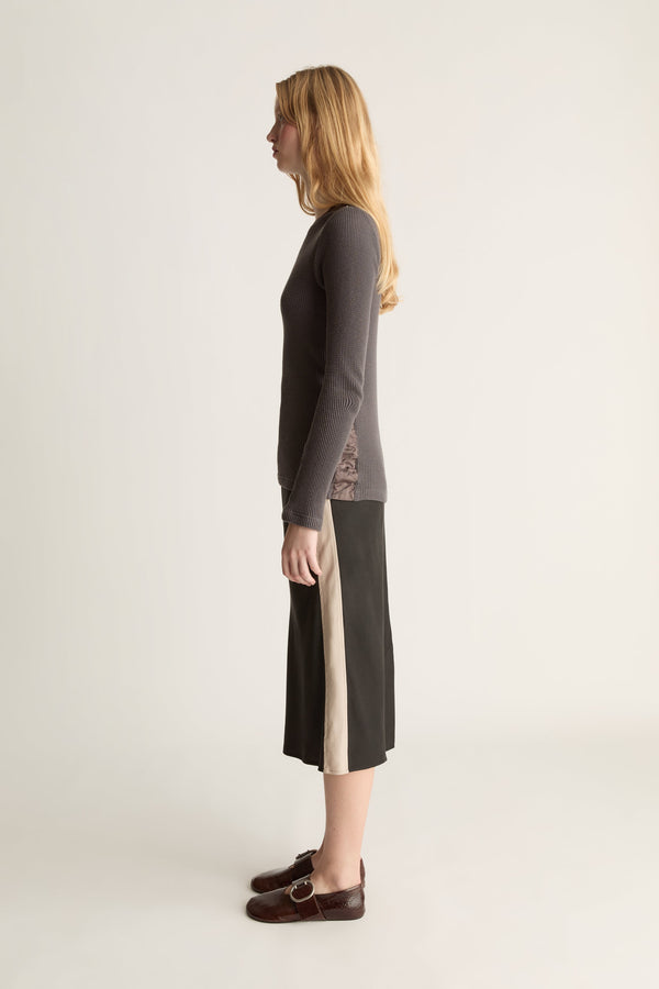A woman stands in profile wearing a dark charcoal long-sleeve knit top and a dark midi skirt with a beige side stripe, and dark brown loafers, against a plain light background.