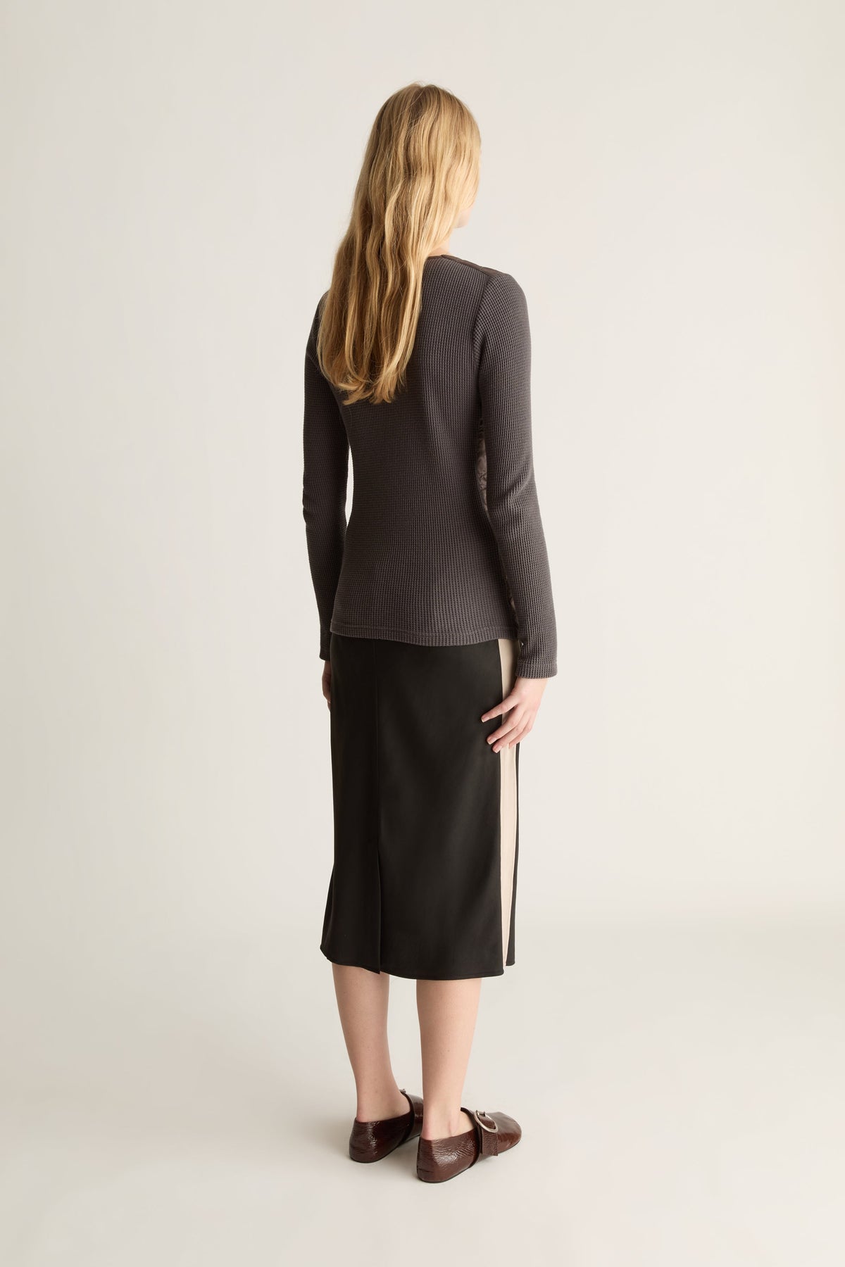 A woman shown from the back wearing a dark charcoal gray long-sleeve knit top and a black midi skirt with a beige side stripe, paired with dark brown loafers, against a light gray studio background.