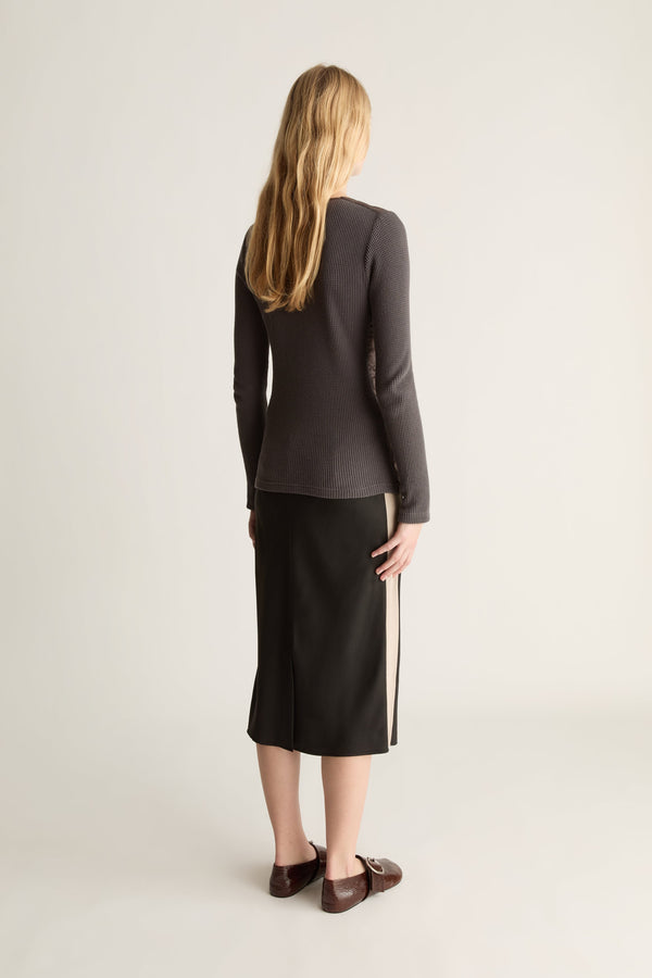 A woman shown from the back wearing a dark charcoal gray long-sleeve knit top and a black midi skirt with a beige side stripe, paired with dark brown loafers, against a light gray studio background.