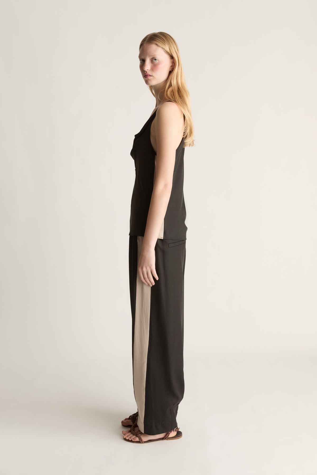 Side view of a woman wearing a sleeveless black blouse and a pair of black trousers with a white stripe down the side on a white background.