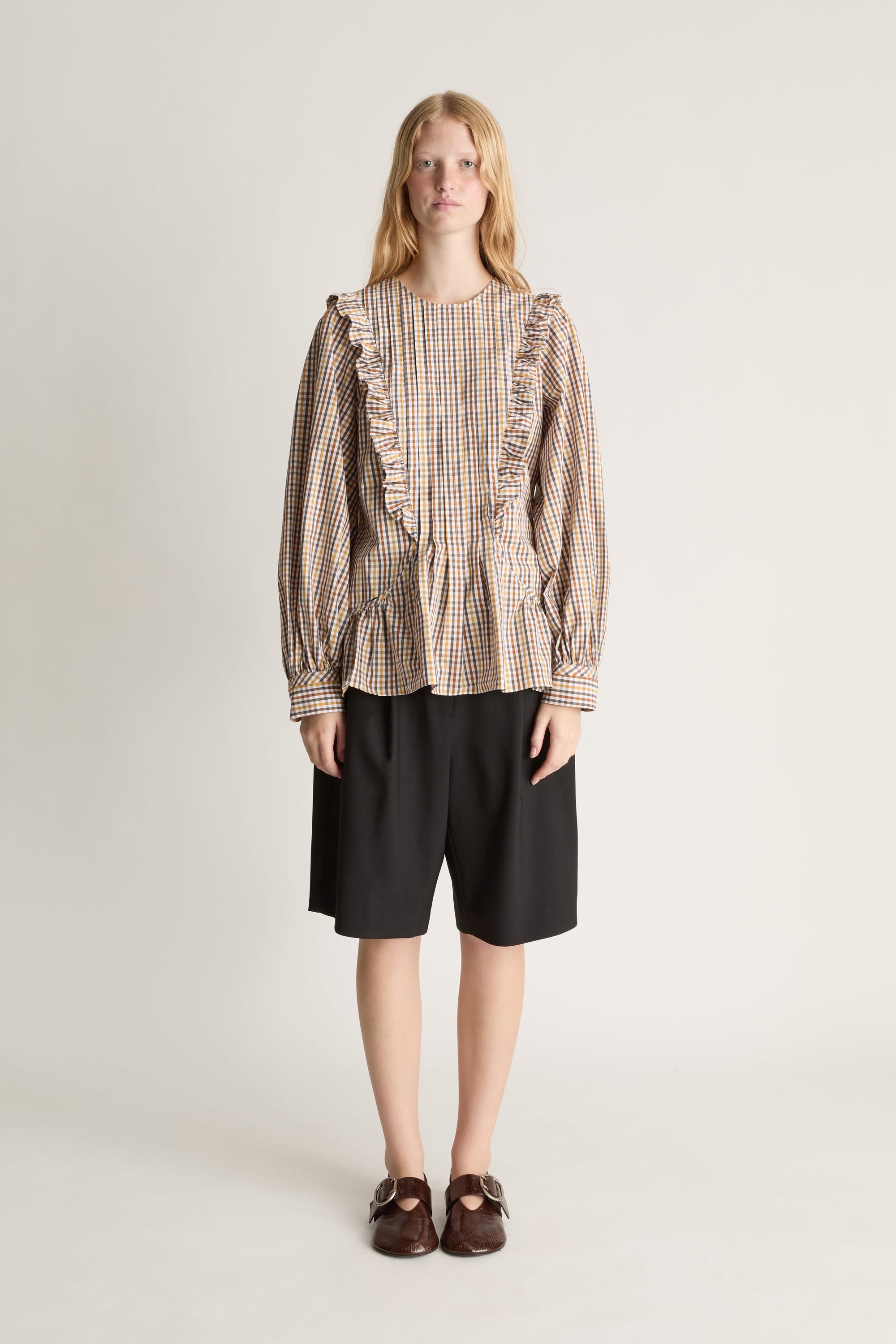 Front view of a person wearing a plaid shirt with ruffles and black shorts on a white background.