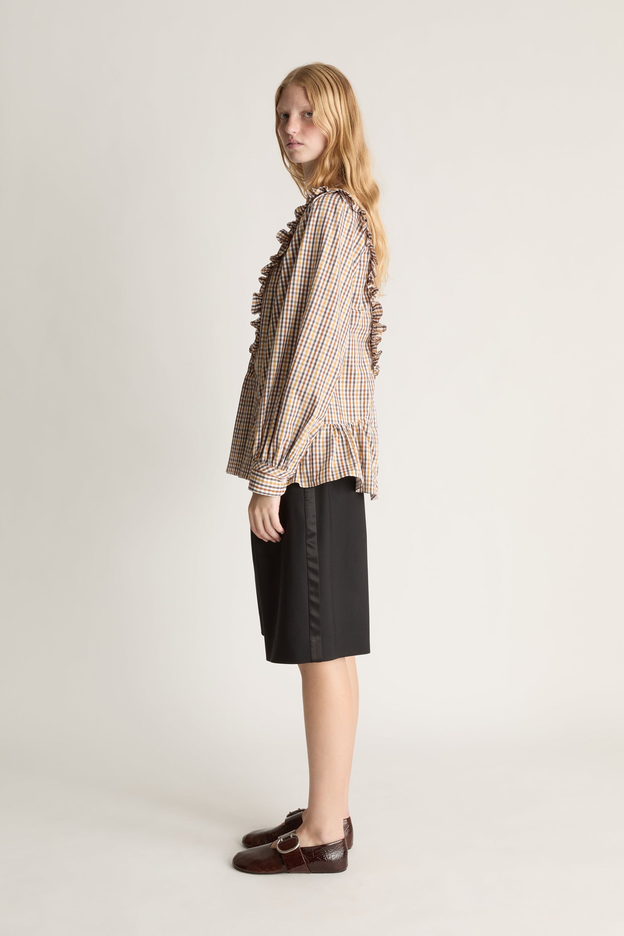 Side view of a woman wearing a plaid long sleeve shirt with ruffles and black shorts on a white background