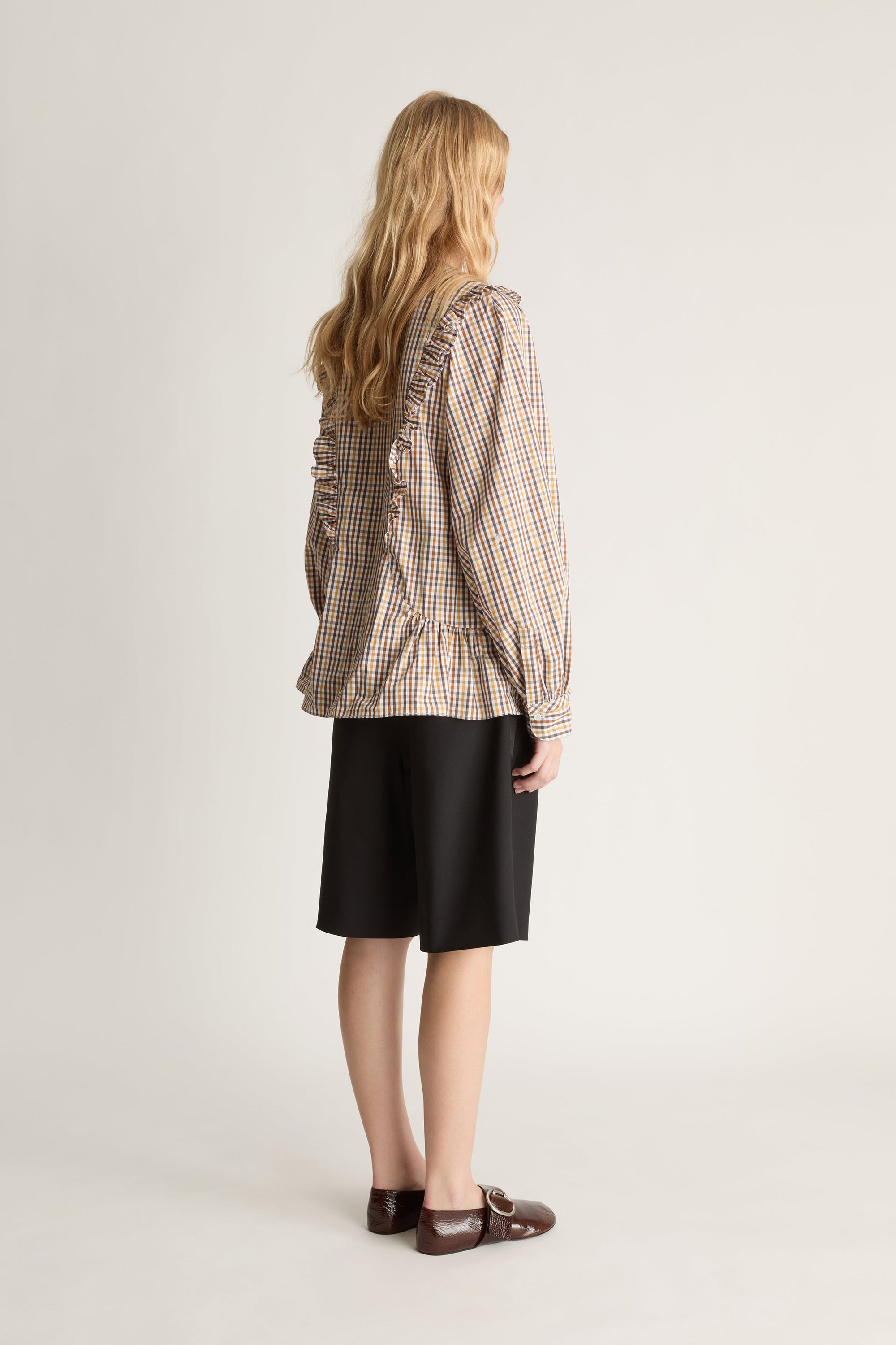 Back view of a woman wearing plaid long sleeve shirt and black shorts against a white studio background.