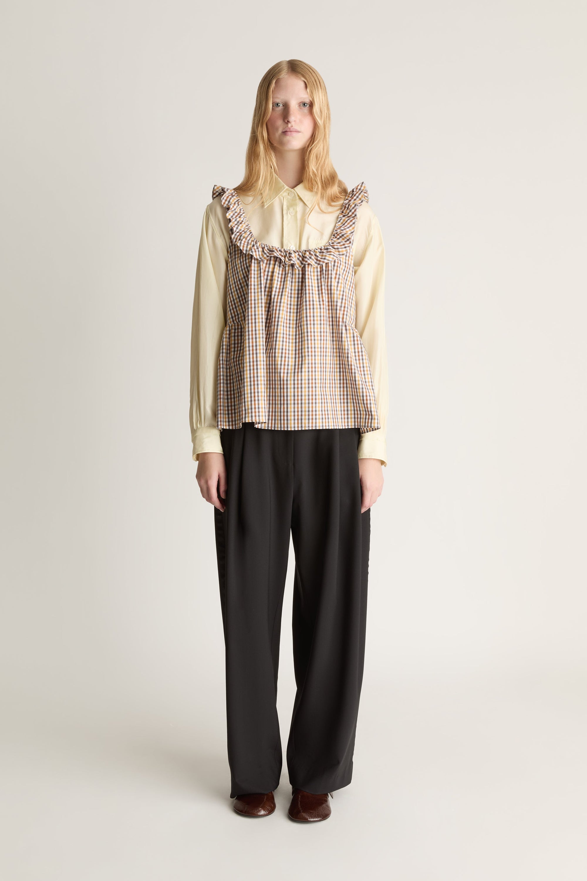 Person wearing a checkered blouse over a yellow long sleeve shirt with ruffled collar and black pants on a white background