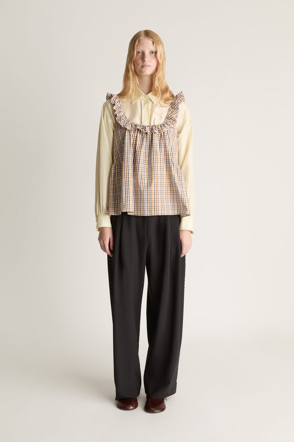 Person wearing a checkered blouse over a yellow long sleeve shirt with ruffled collar and black pants on a white background