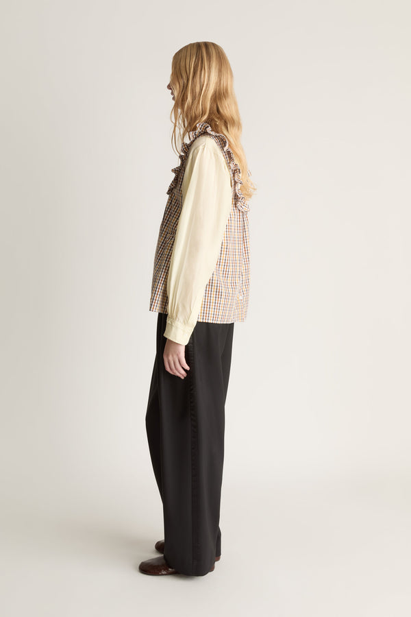Profile view of a woman wearing a check blouse over a yellow long sleeve shirt with black trousers against a white studio bakground. 