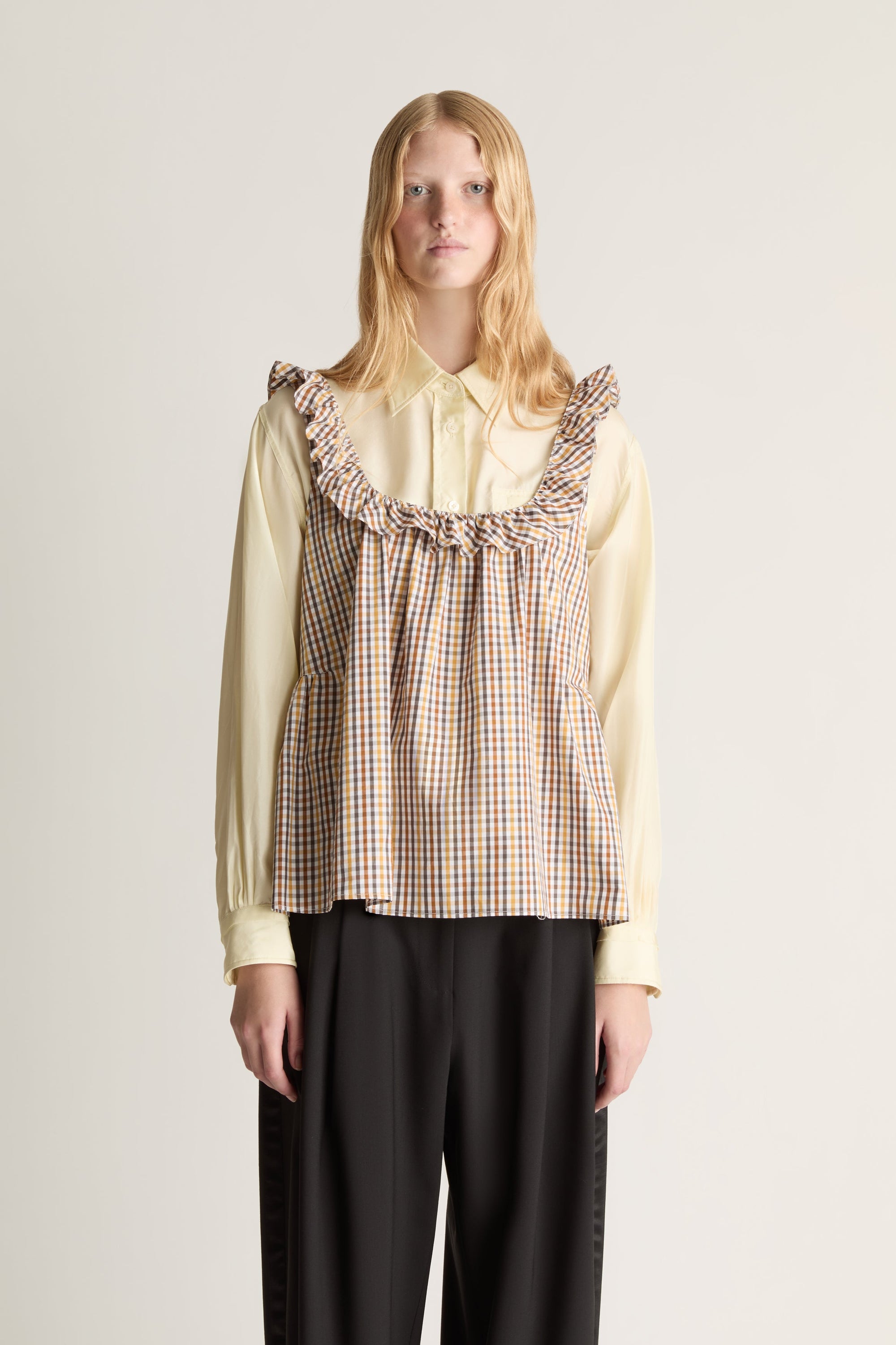 Close up of a woman wearing a checkered blouse over a yellow shirt with ruffled details on a plain background