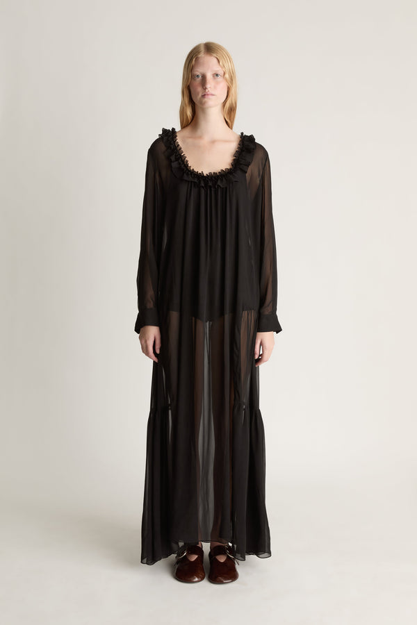 Front view of a woman wearing a black long-sleeve sheer dress with a frill neckline against a white studio background. 