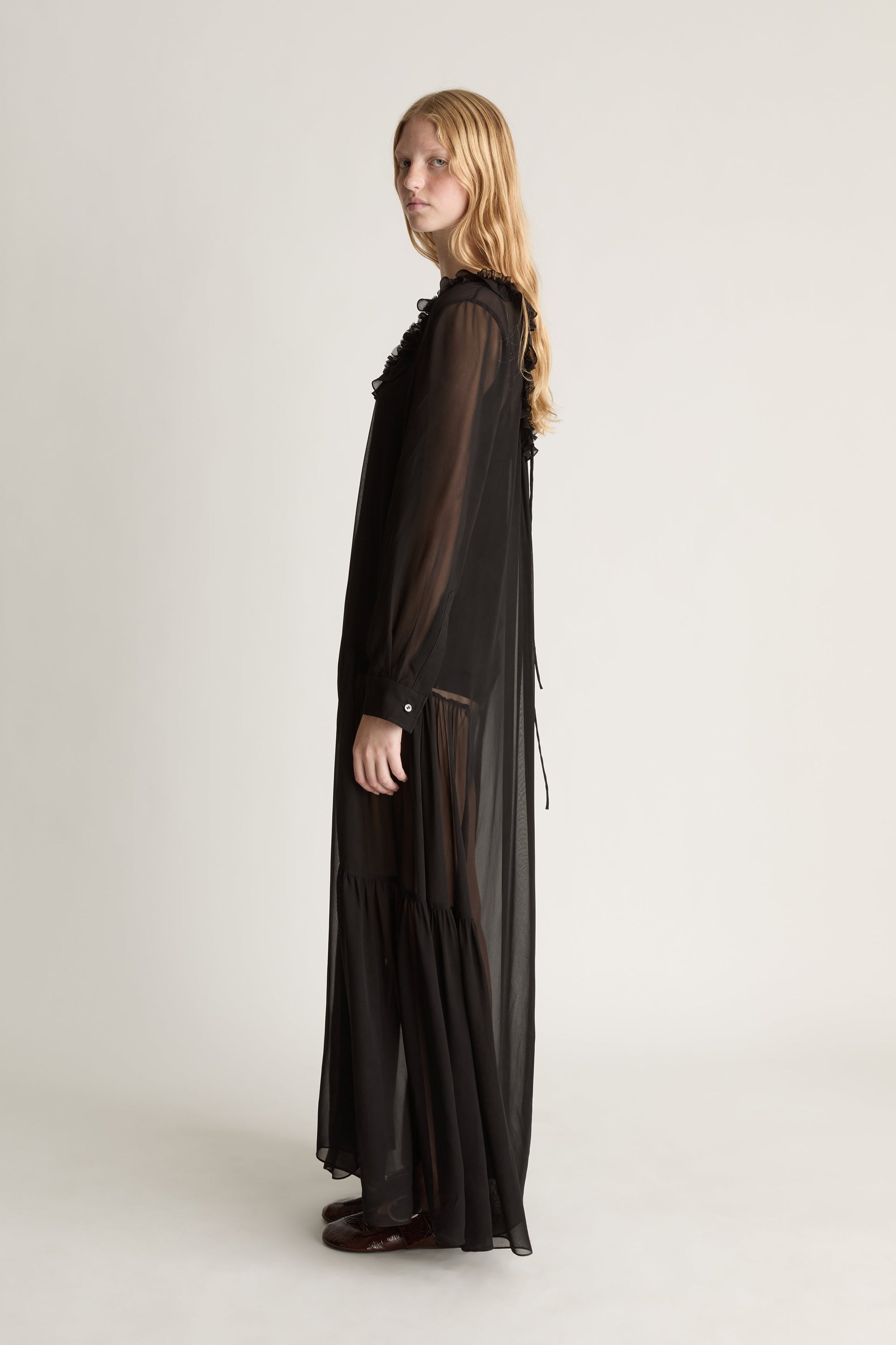 Profile view of a woman wearing a back long sleeve sheer dress agaisnt a white background. 