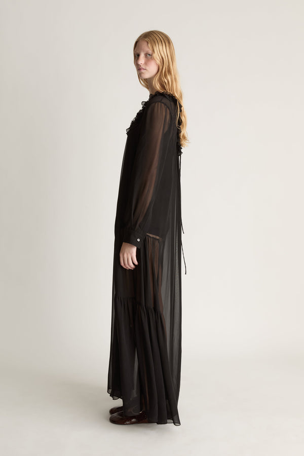 Profile view of a woman wearing a back long sleeve sheer dress agaisnt a white background. 