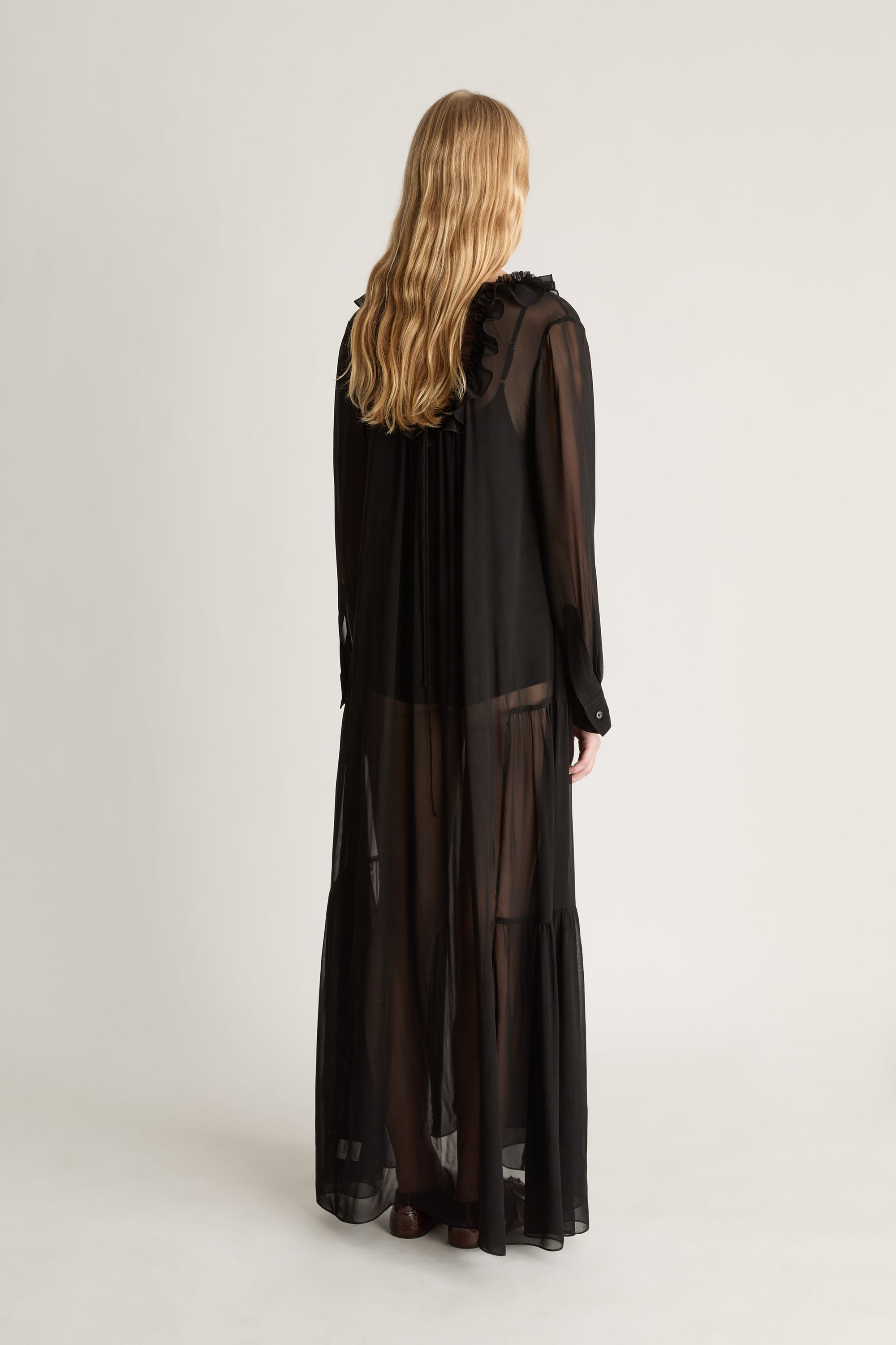 Back view of a woman wearing a long sleeve, black sheer georgette dress against a white studio background. 