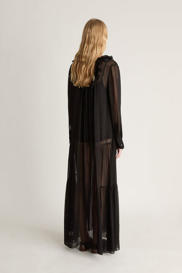 Back view of a woman wearing a long sleeve, black sheer georgette dress against a white studio background. 