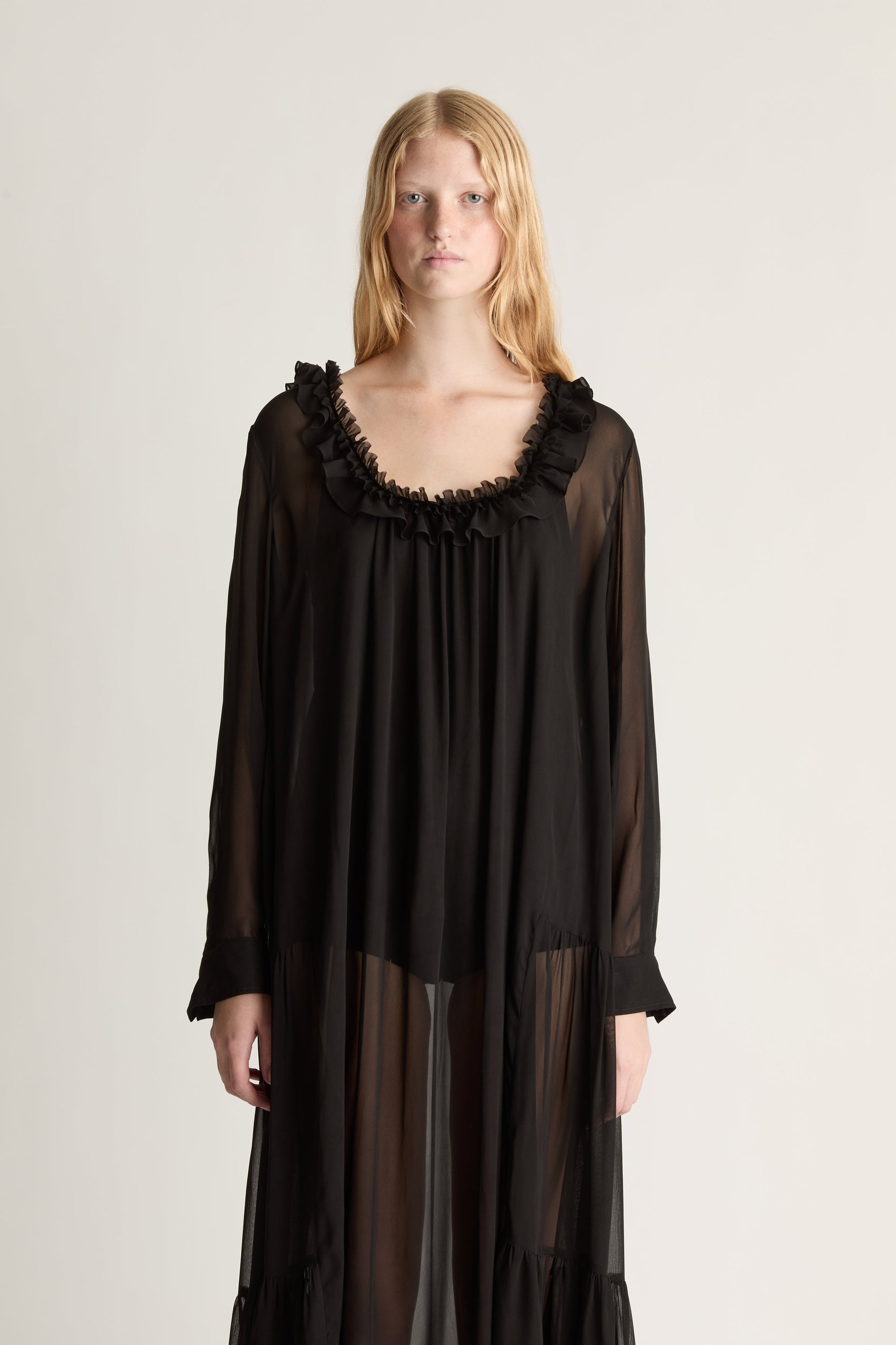 Close up of a woman wearing a black long sleeve sheer georgette dress against a white studio background.