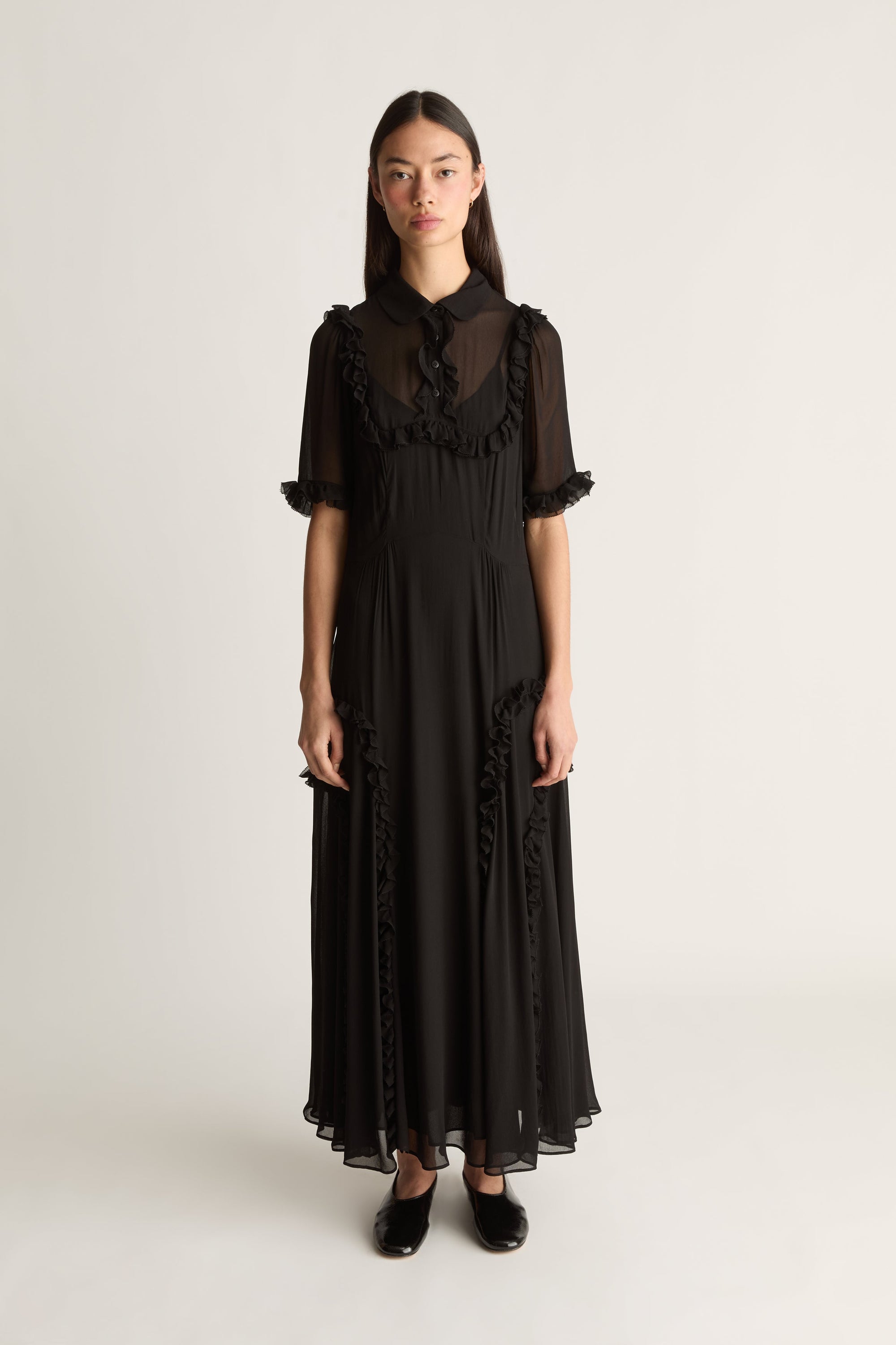 A model wearing a black midi dress with sheer short sleeves, a high collar, and ruffled detailing, standing against a plain light gray studio background.