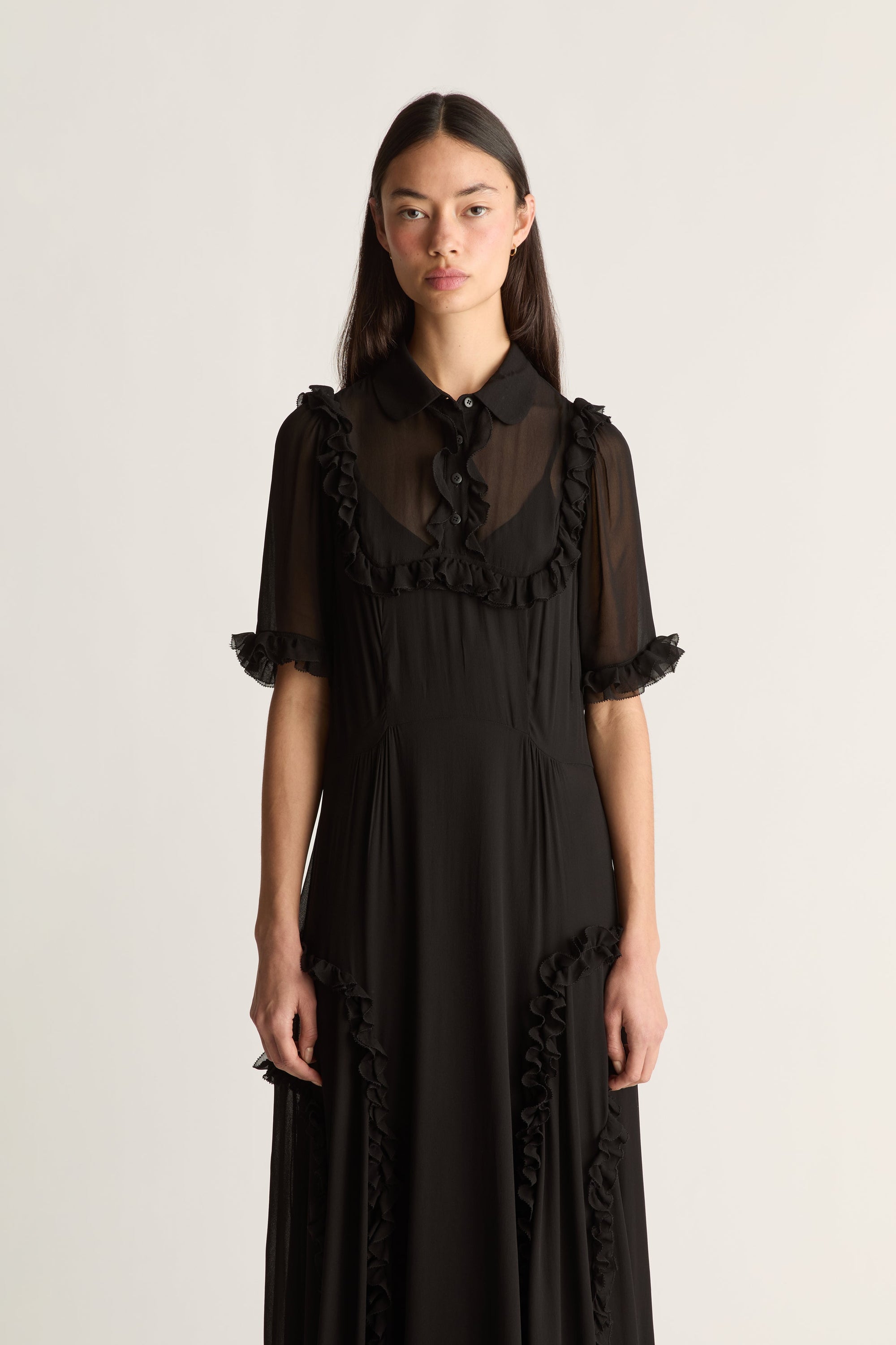 A model wearing a black midi dress with sheer ruffled short sleeves, a ruffled collar, and ruffle trim along the front and sides, photographed against a plain light background.