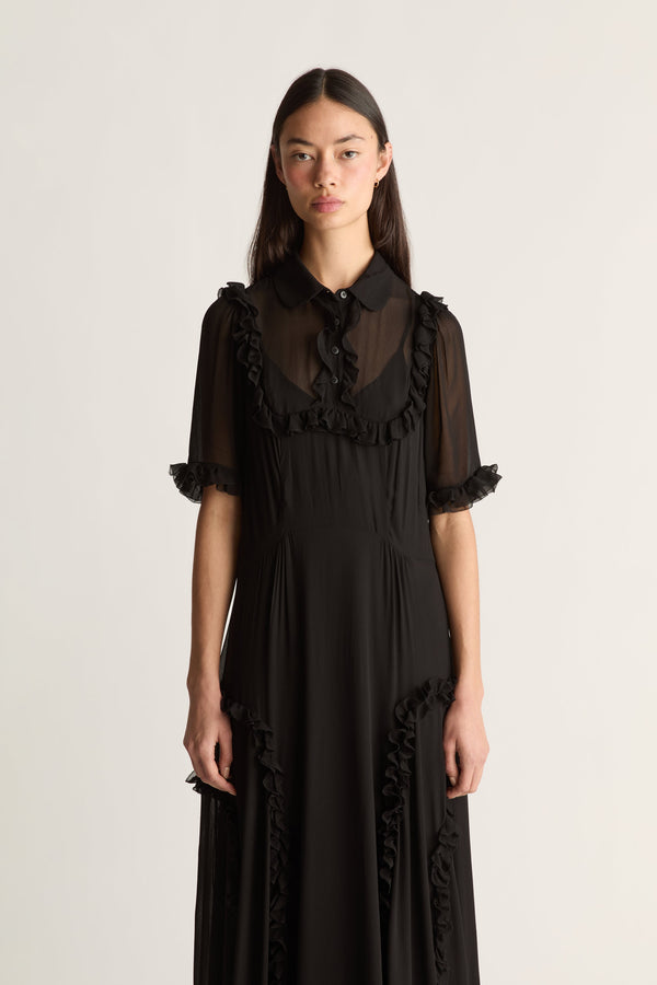 A model wearing a black midi dress with sheer ruffled short sleeves, a ruffled collar, and ruffle trim along the front and sides, photographed against a plain light background.