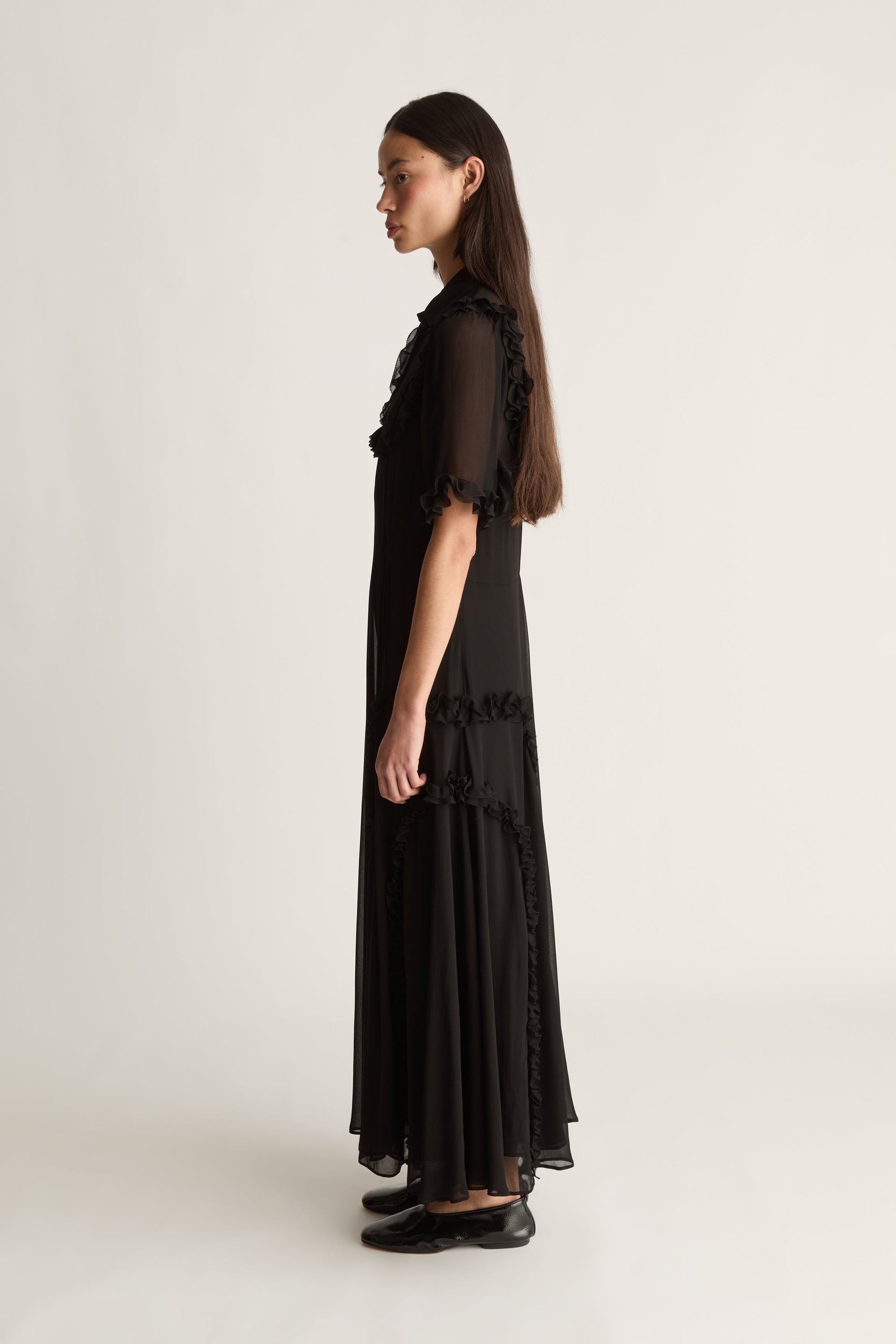 A woman stands in profile wearing a long black dress with sheer ruffled short sleeves and a flowing skirt, photographed against a plain light gray studio background.
