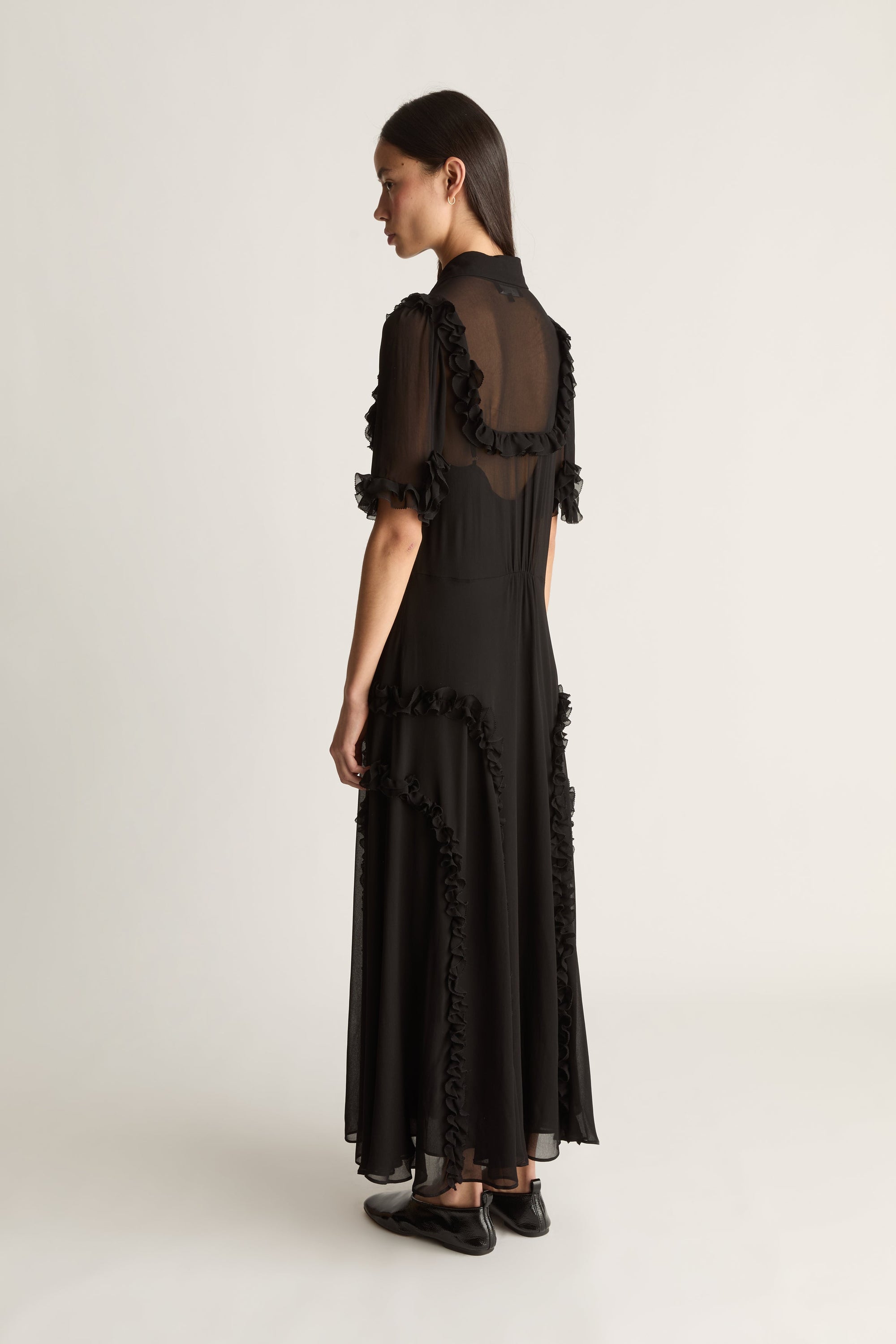 A model is shown from the back wearing a long black dress with sheer ruffled short sleeves and a high neckline, paired with black shoes, against a plain light gray studio background.