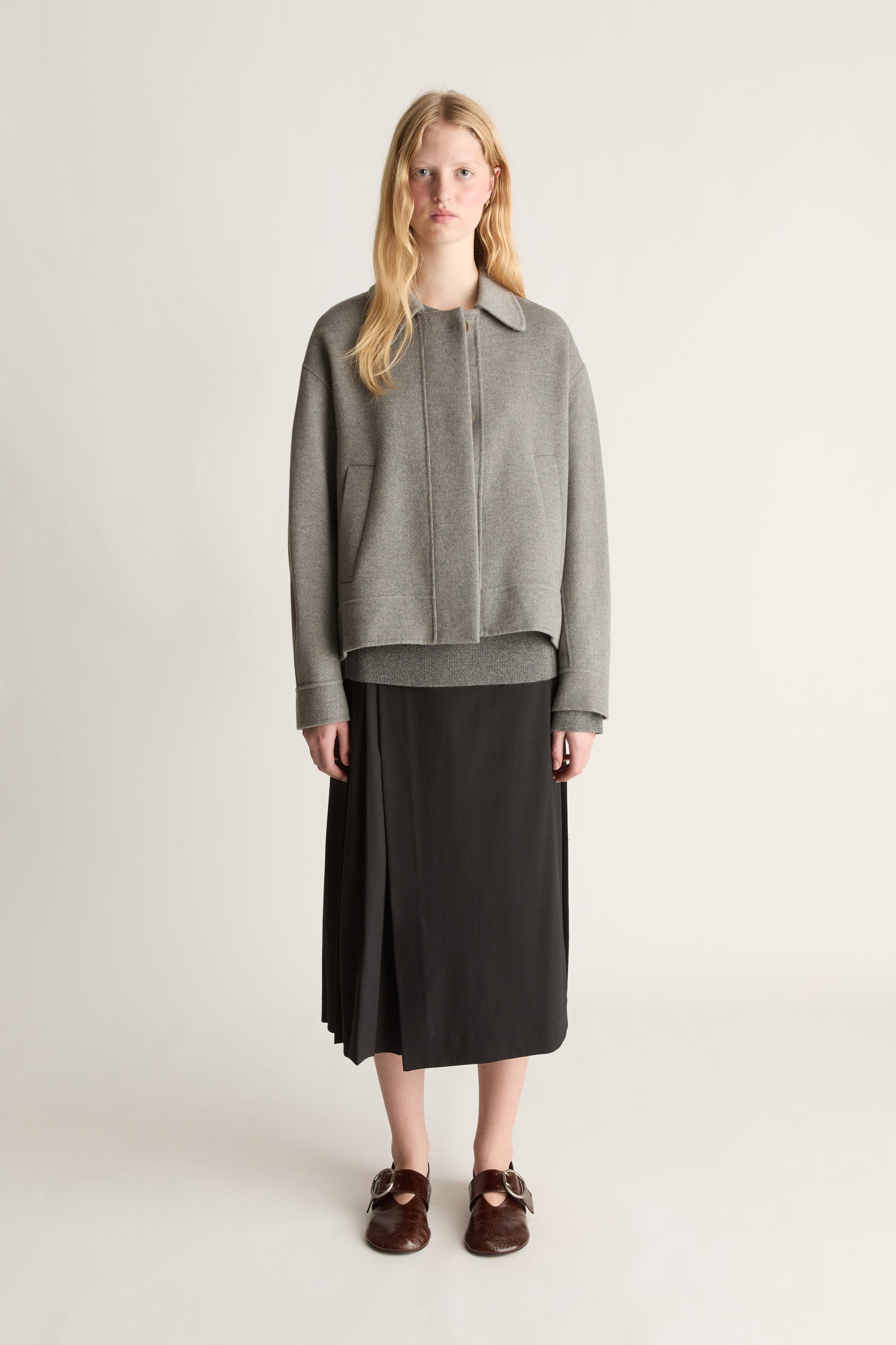 Front view of a woman wearing a grey cashmere worker jacket and a black kilt skirt with brown shoes against a white background.