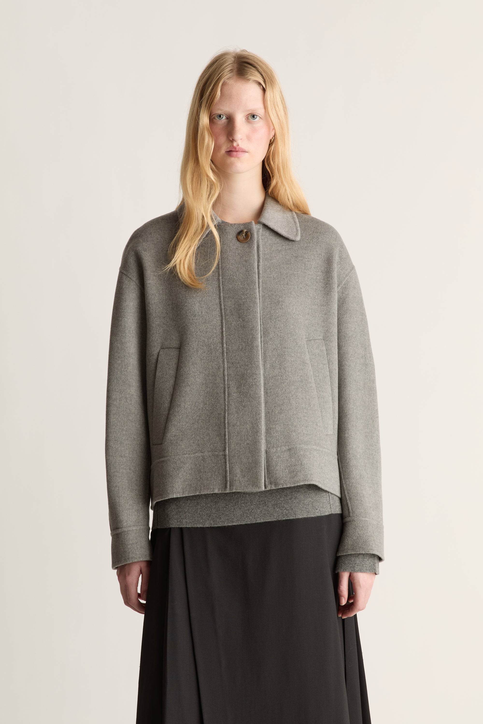 Close up of a woman wearing a light grey worker jacket and a black skirt against a white studio background.