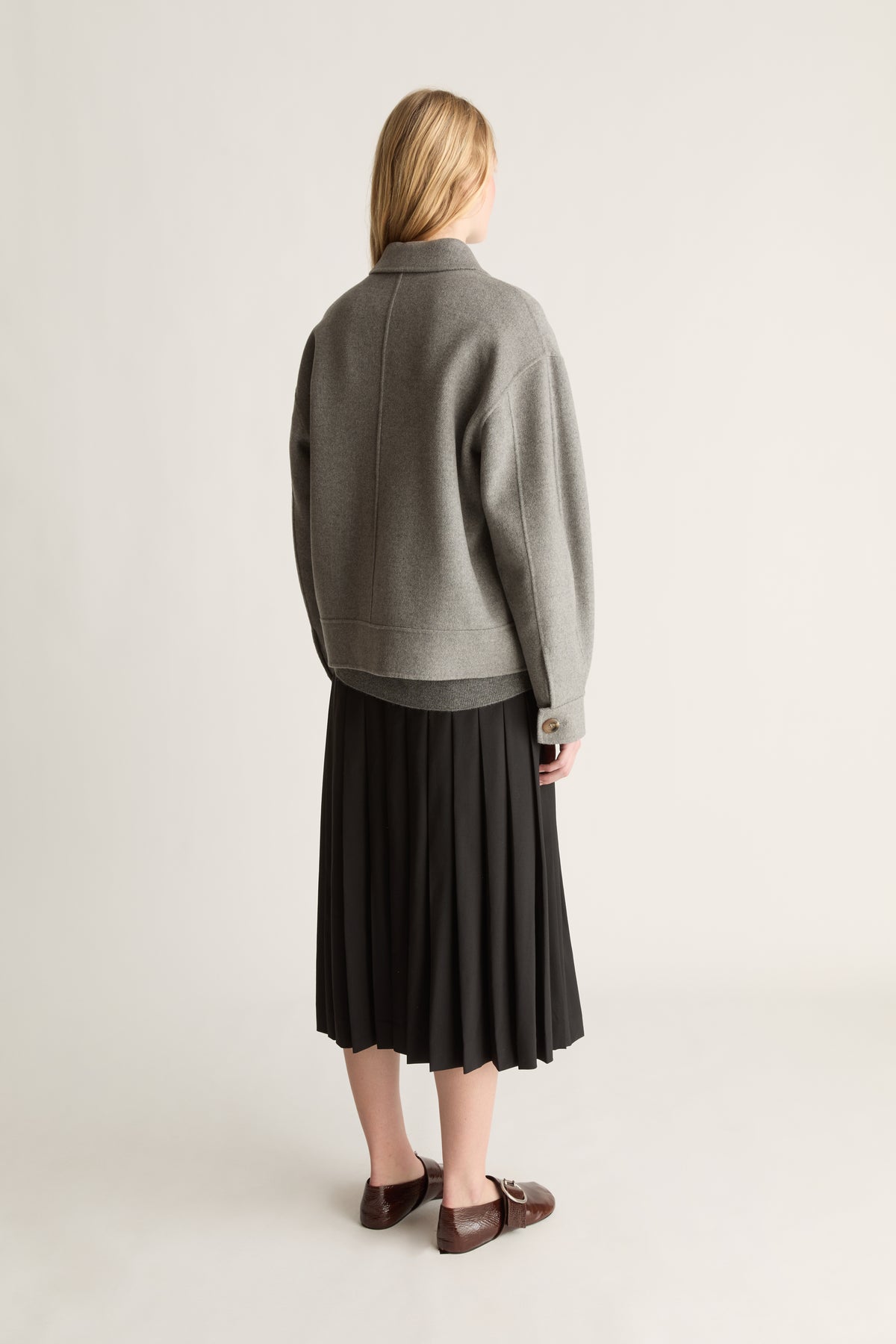 Back view of a woman wearing a grey jacket, a black wool kilt skirt and brown shoes against a white studio background..