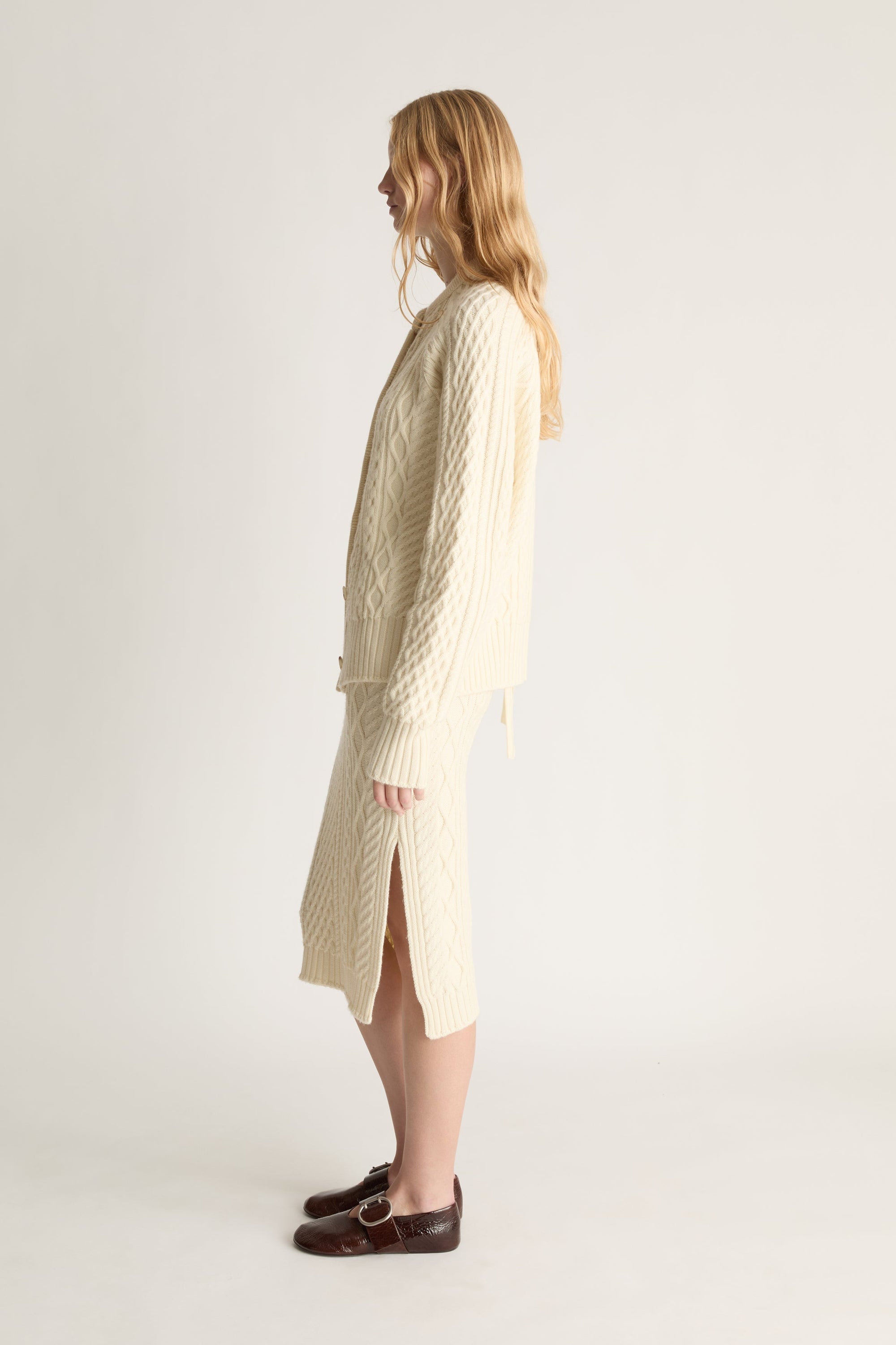 A woman stands in profile wearing a cream cable-knit cardigan and matching midi dress, paired with dark brown loafers, against a plain light gray studio background.