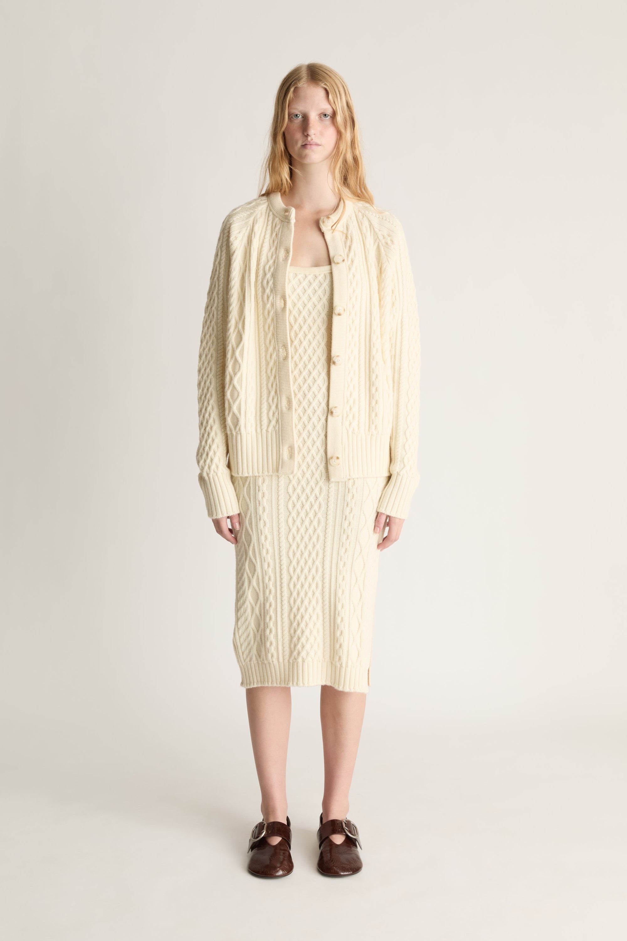 Woman wearing a cable knit midi dress and a matching cardigan against a white studio background. 