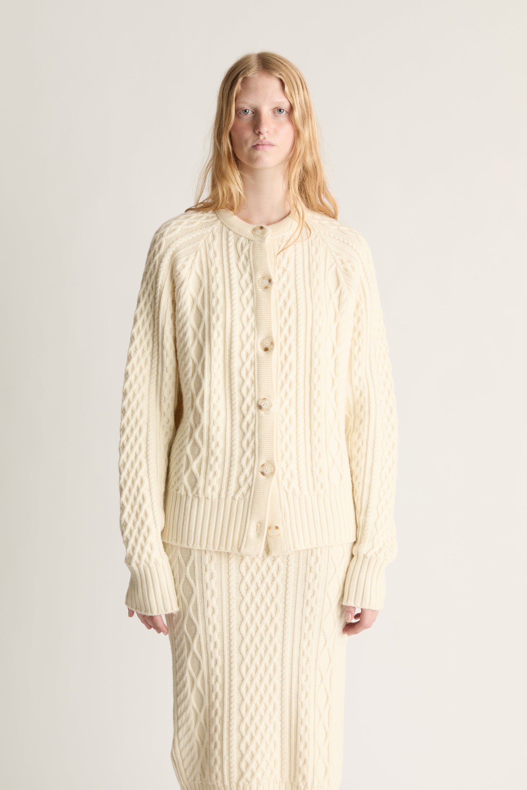 Woman wearing a cream-colored cable knit cardigan and dress set against a plain background